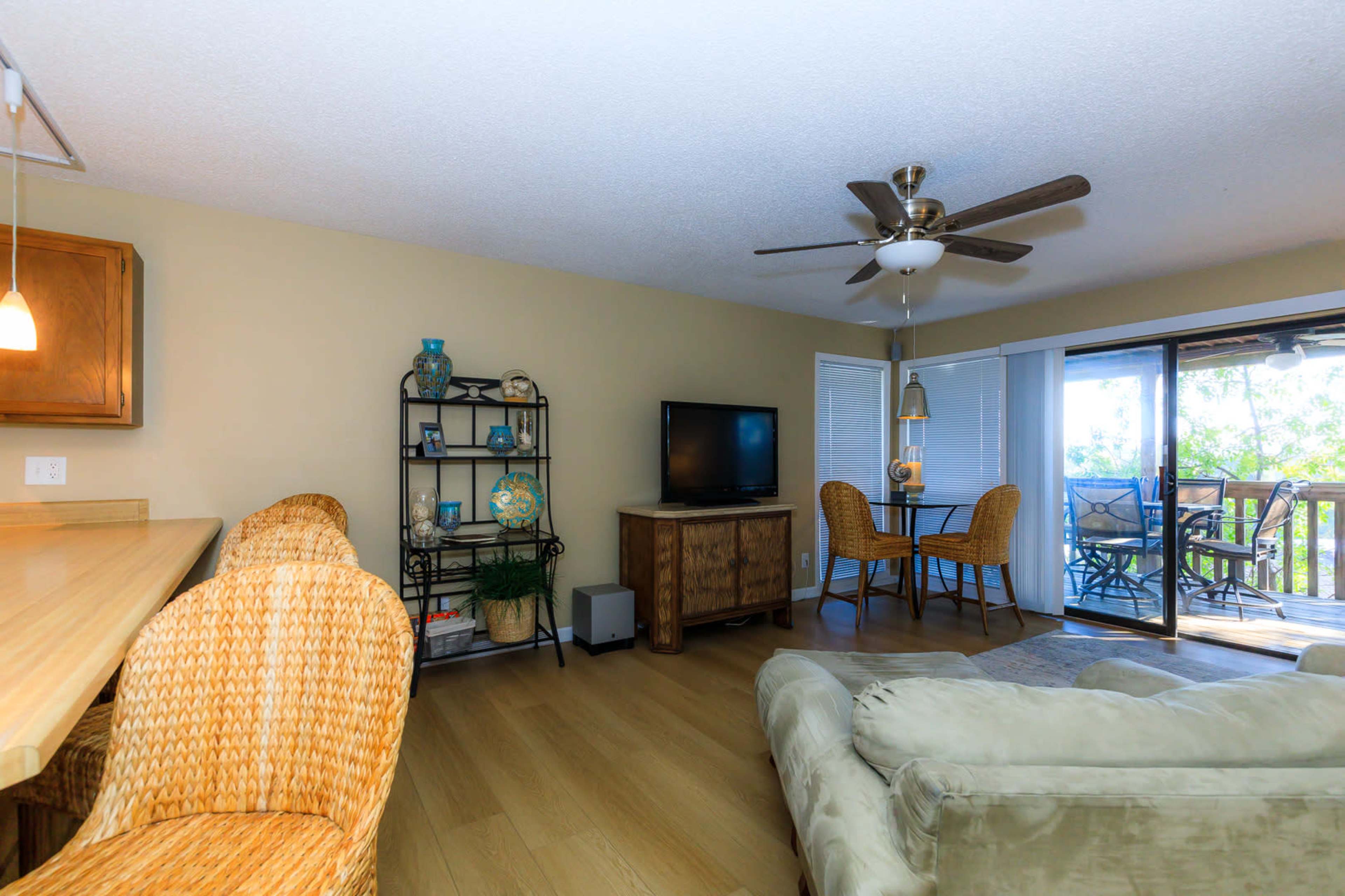 A cozy living area with a beige sofa, a television on a wooden stand, a dining table for four, and sliding glass doors leading to a balcony.