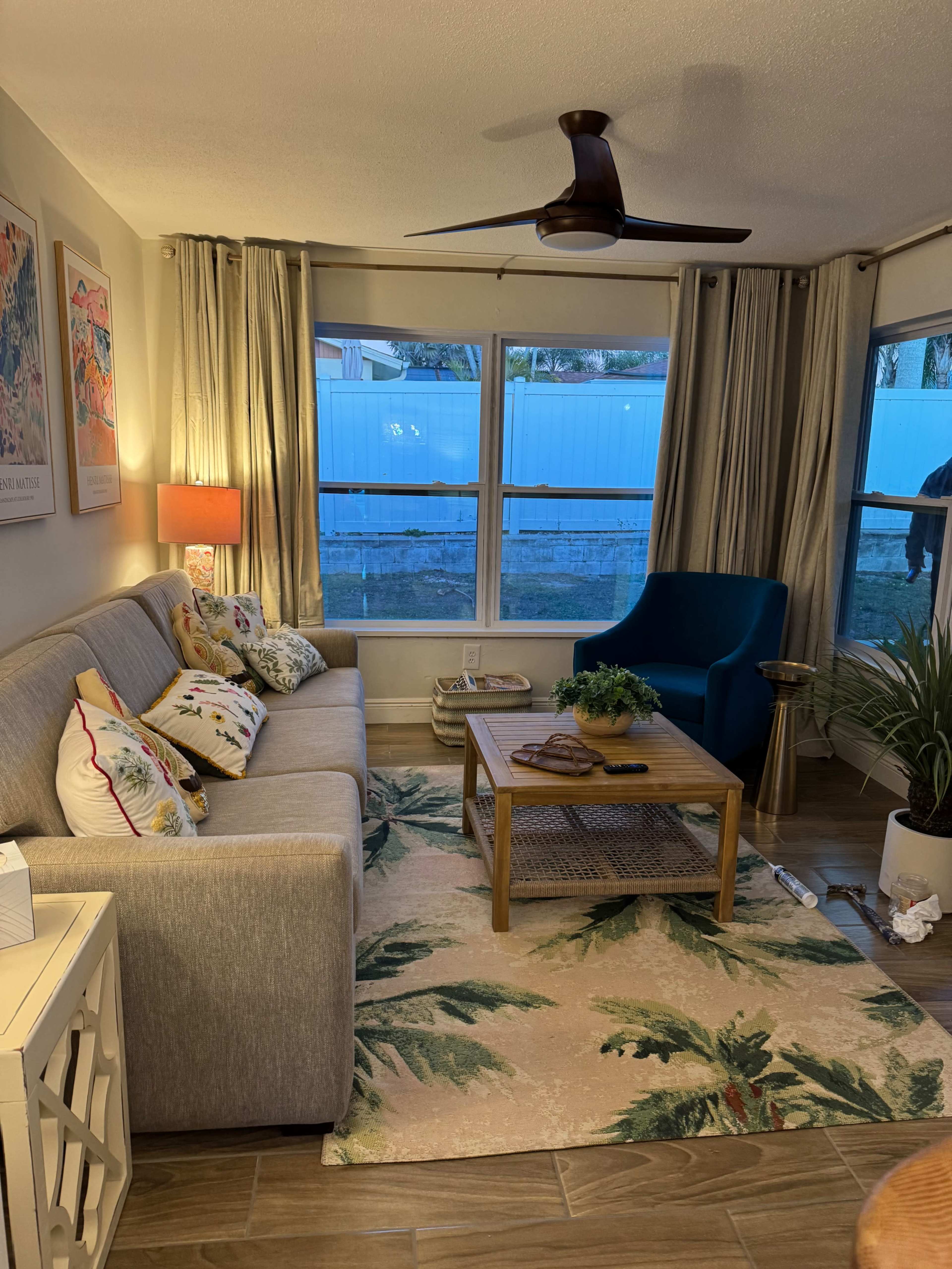 The living room features a gray sofa with patterned pillows, a wooden coffee table, a palm-patterned area rug, and a blue armchair, with large windows letting in natural light.