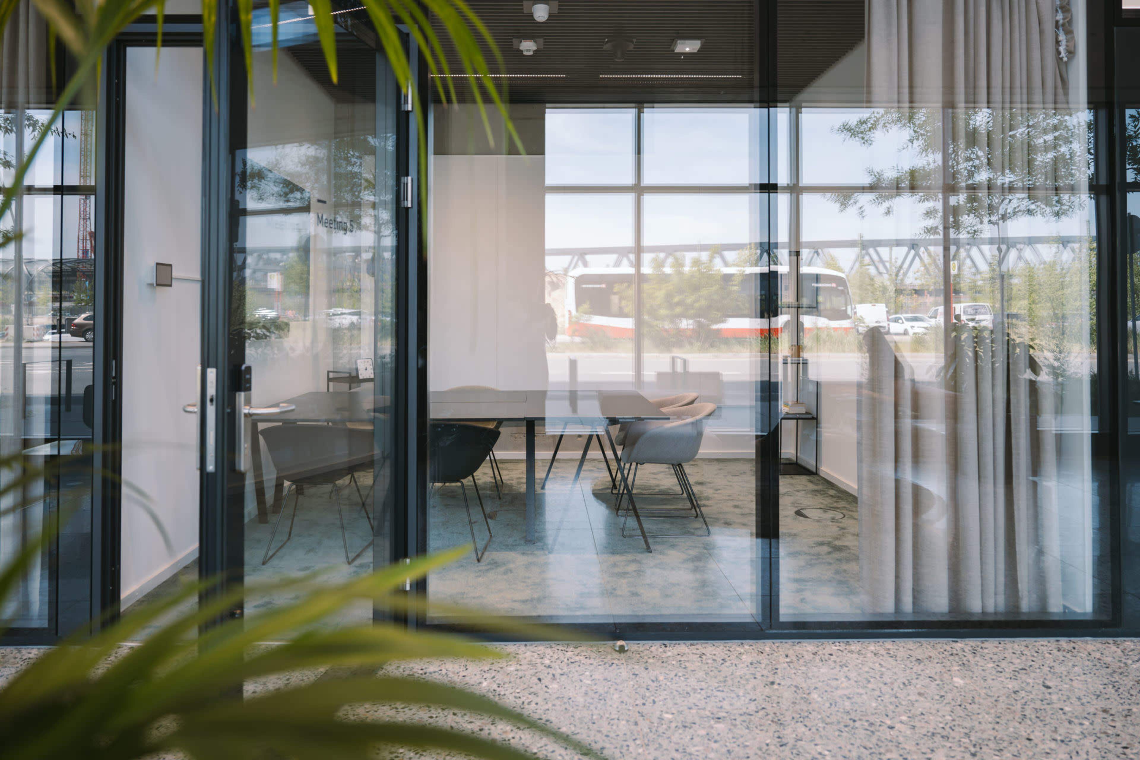 The image shows a modern glass-walled meeting room with tables and chairs, partially obscured by indoor plants.
