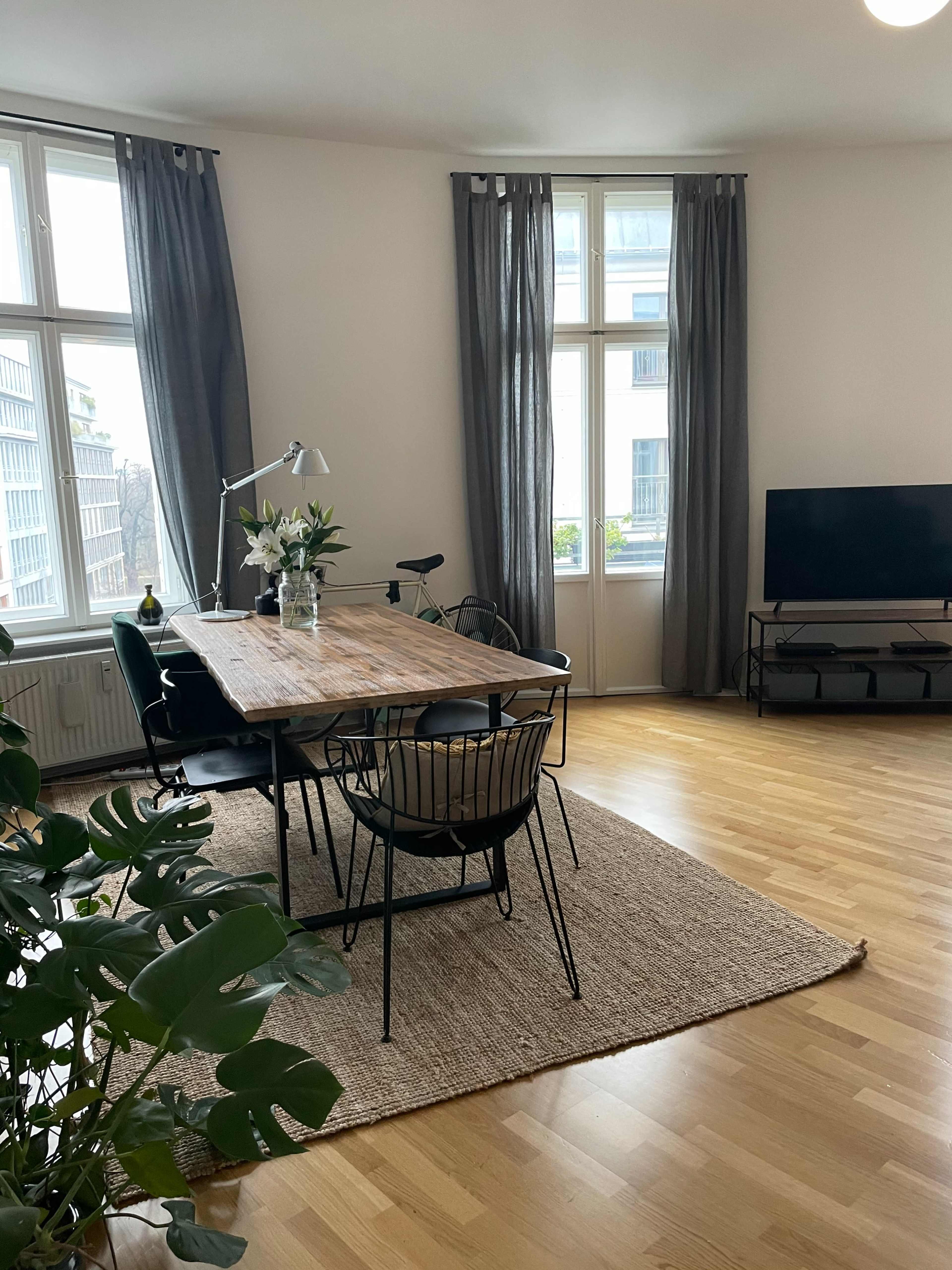 The image shows a modern living and dining area featuring a wooden table surrounded by black chairs, large windows with gray curtains, and a television on a stand.