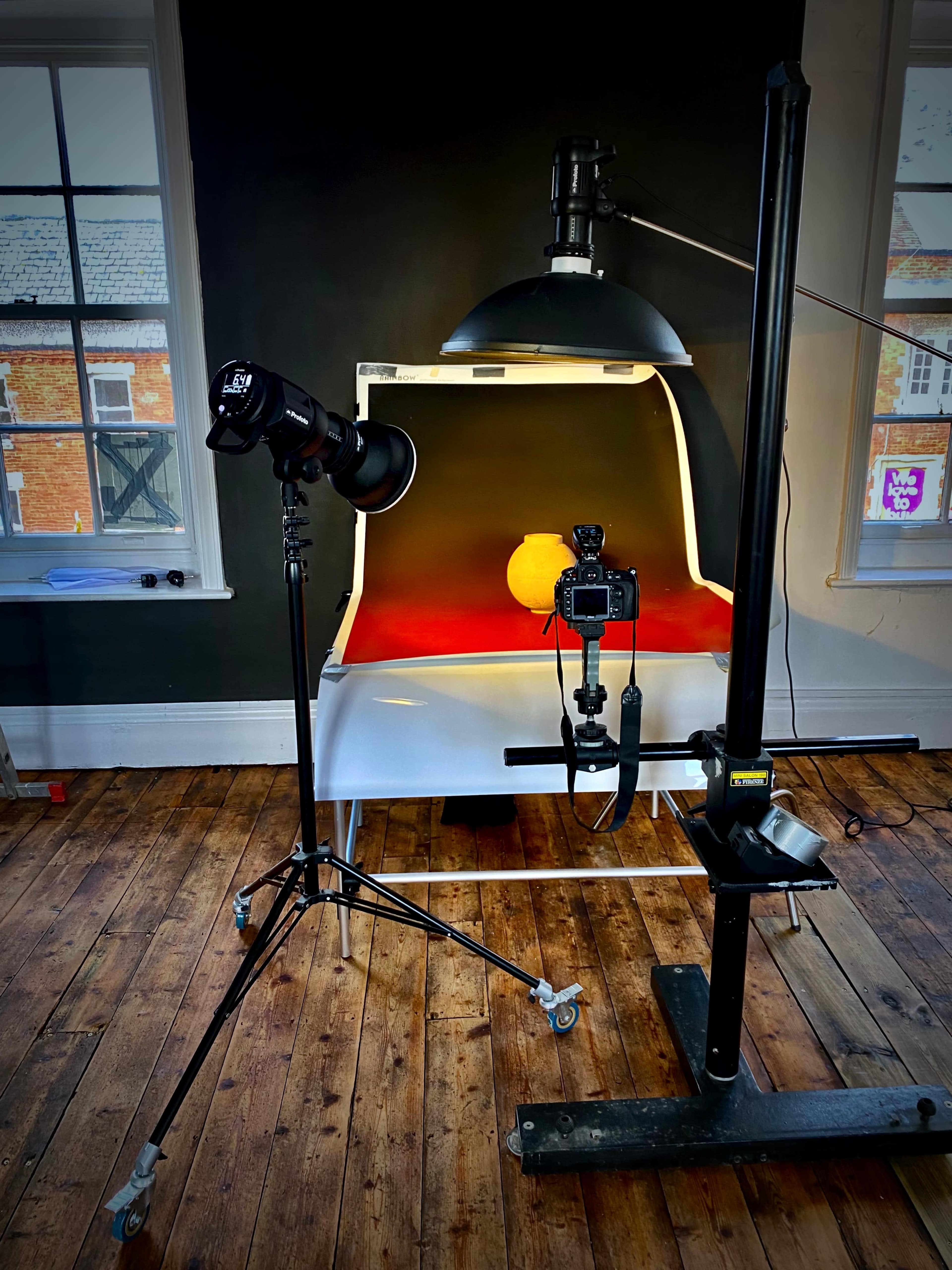 A photography setup features a camera and light positioned in front of a curved white backdrop, illuminating a round object on a red surface in a room with wooden floors.