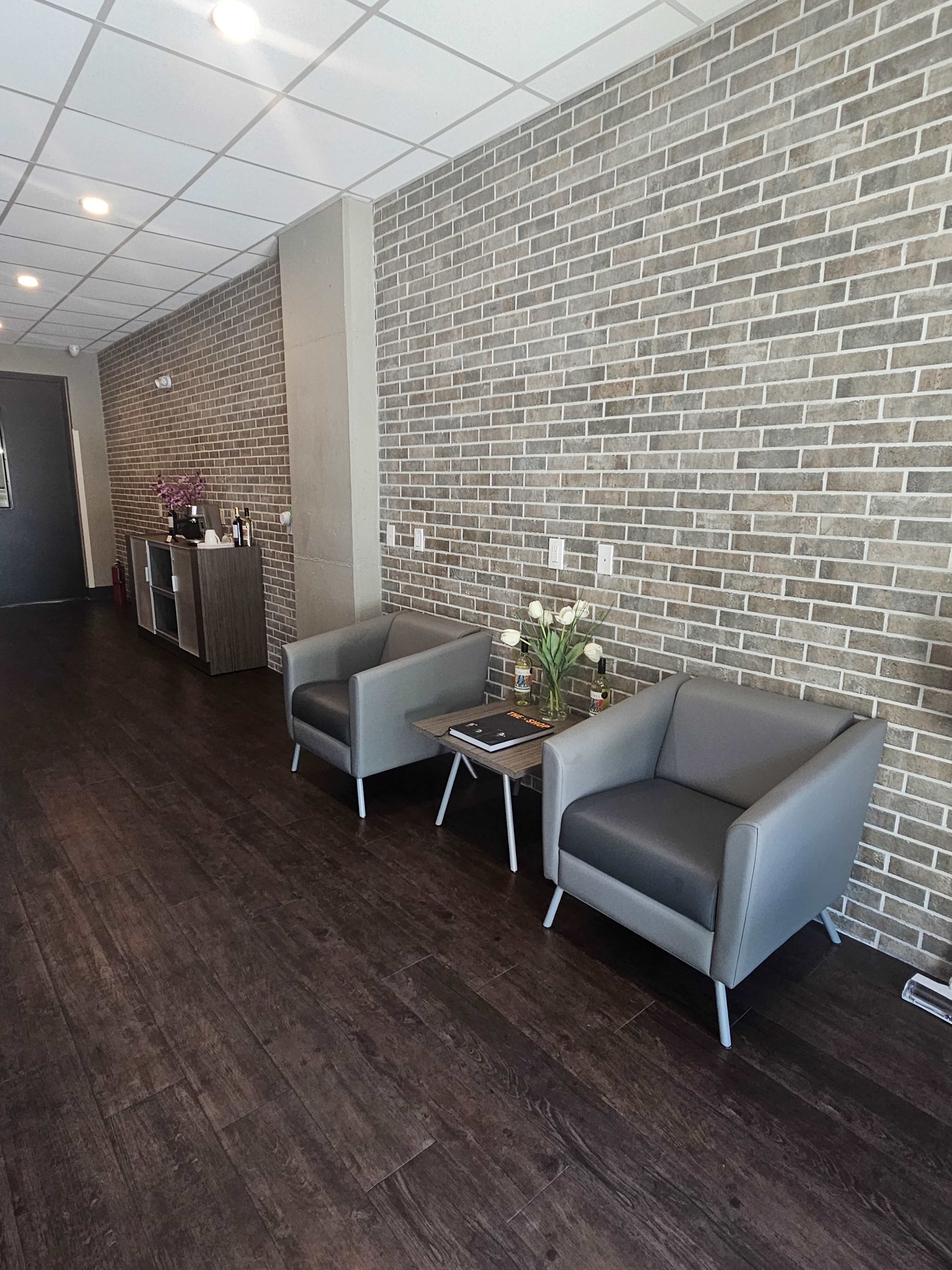 The image shows a modern waiting area featuring two gray chairs, a small table with items on it, and a brick accent wall.