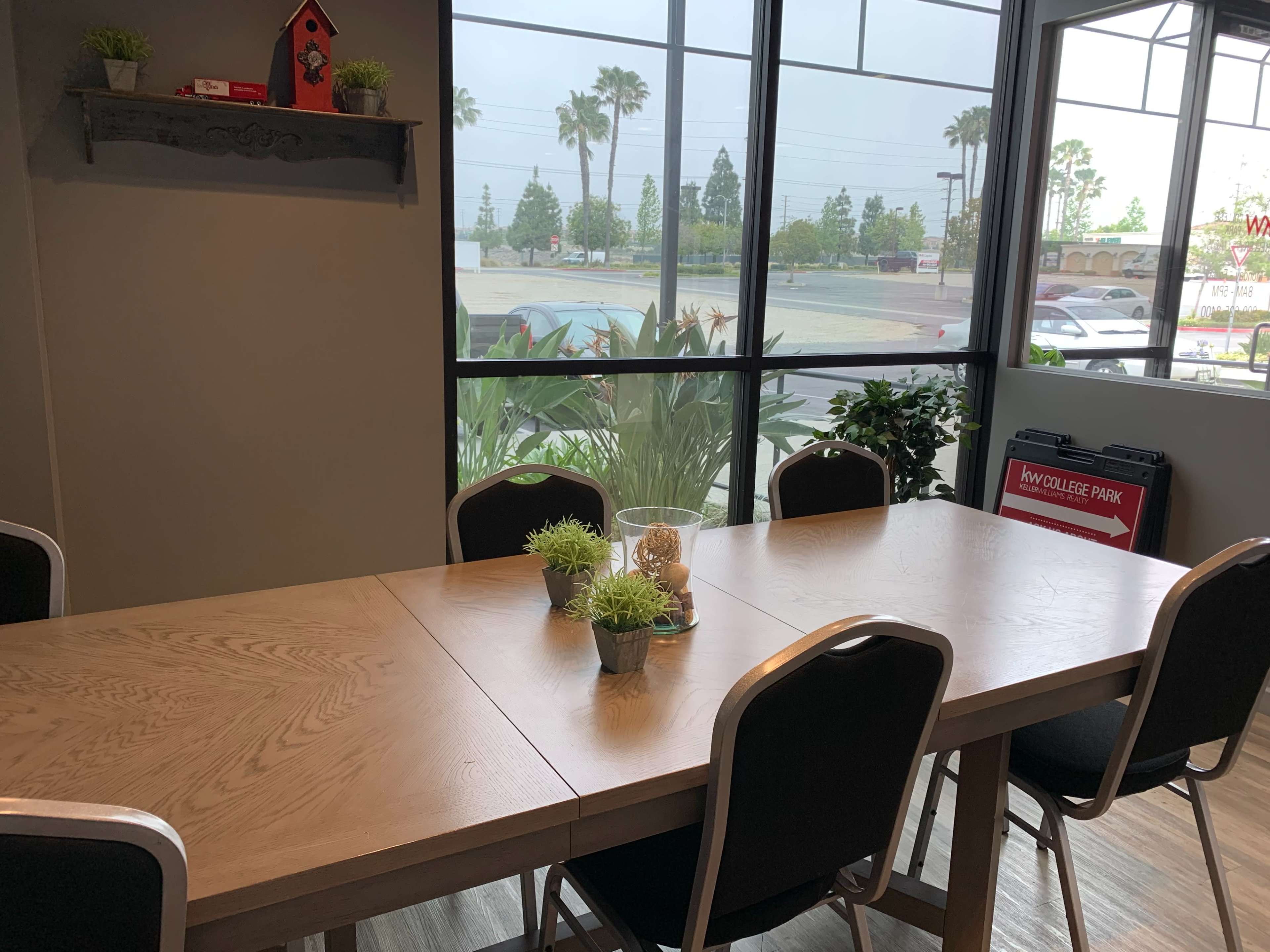 A wooden dining table with small potted plants in the center is set in a well-lit room with large windows overlooking a parking lot and palm trees.