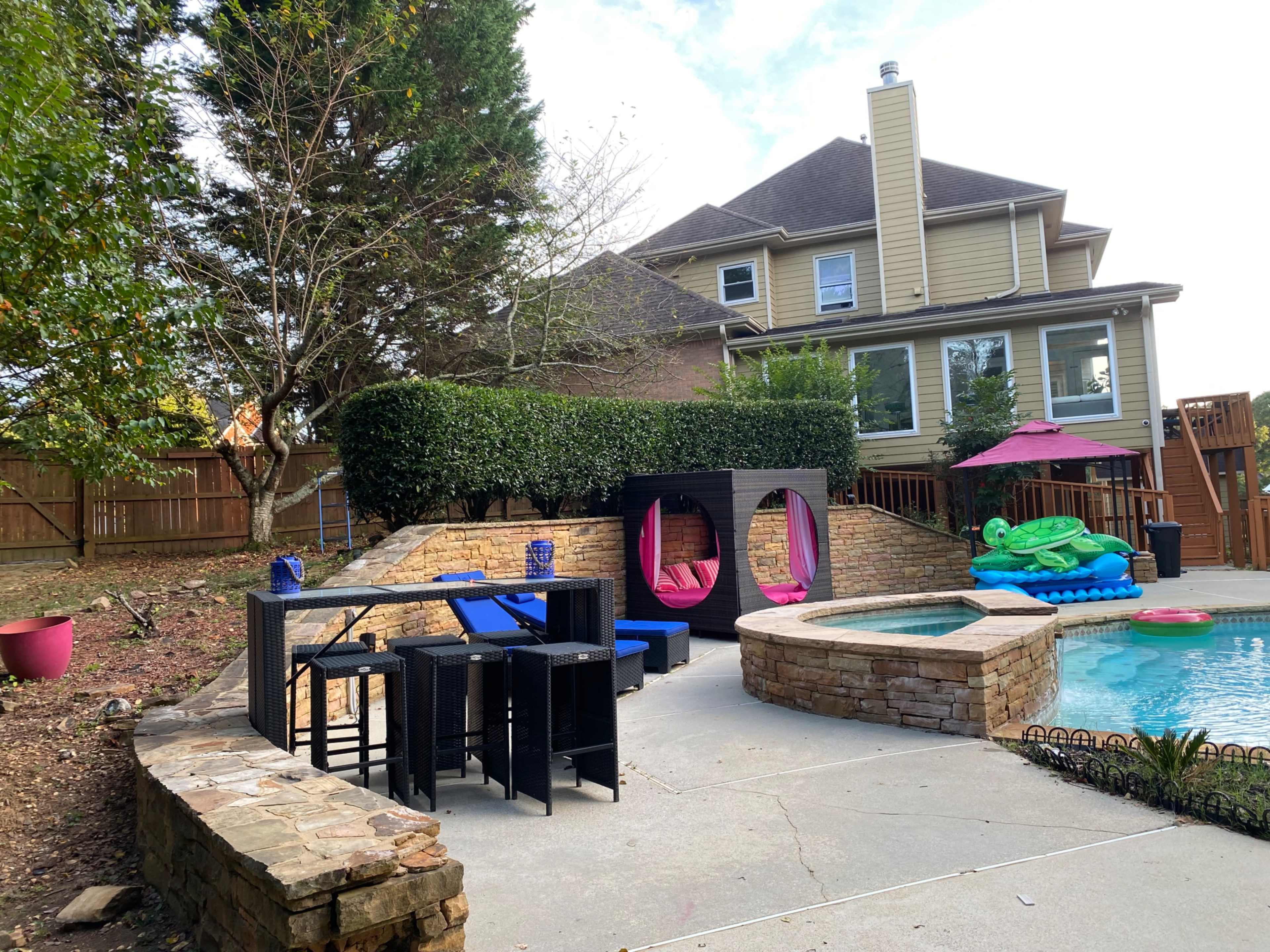 The image shows a backyard with a stone patio, a swimming pool, and outdoor furniture including chairs and a table, surrounded by greenery and a two-story house.