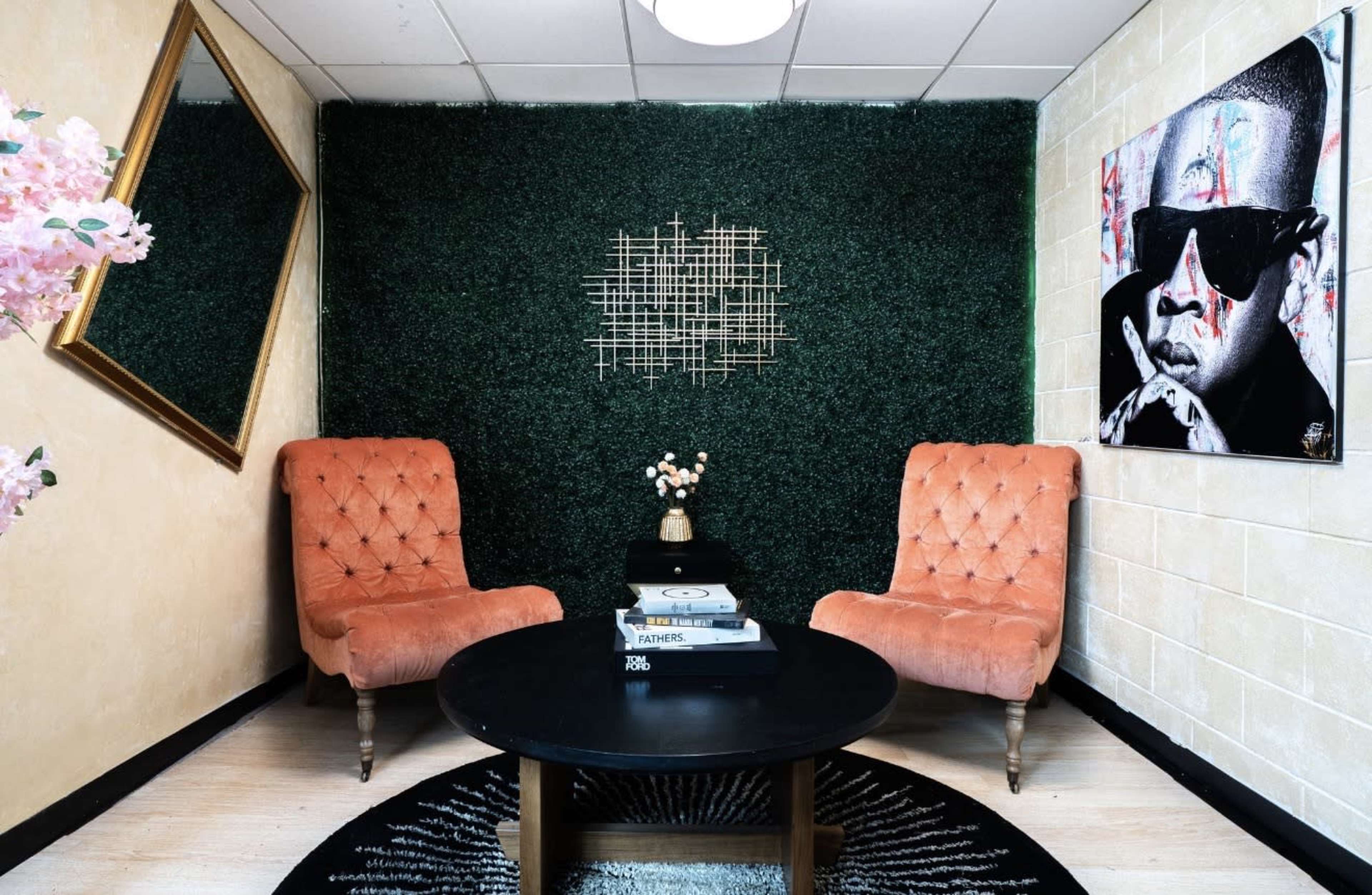 The image shows a small room with two orange armchairs facing a round black coffee table, surrounded by a green hedge wall, a framed artwork, and a decorative mirror.