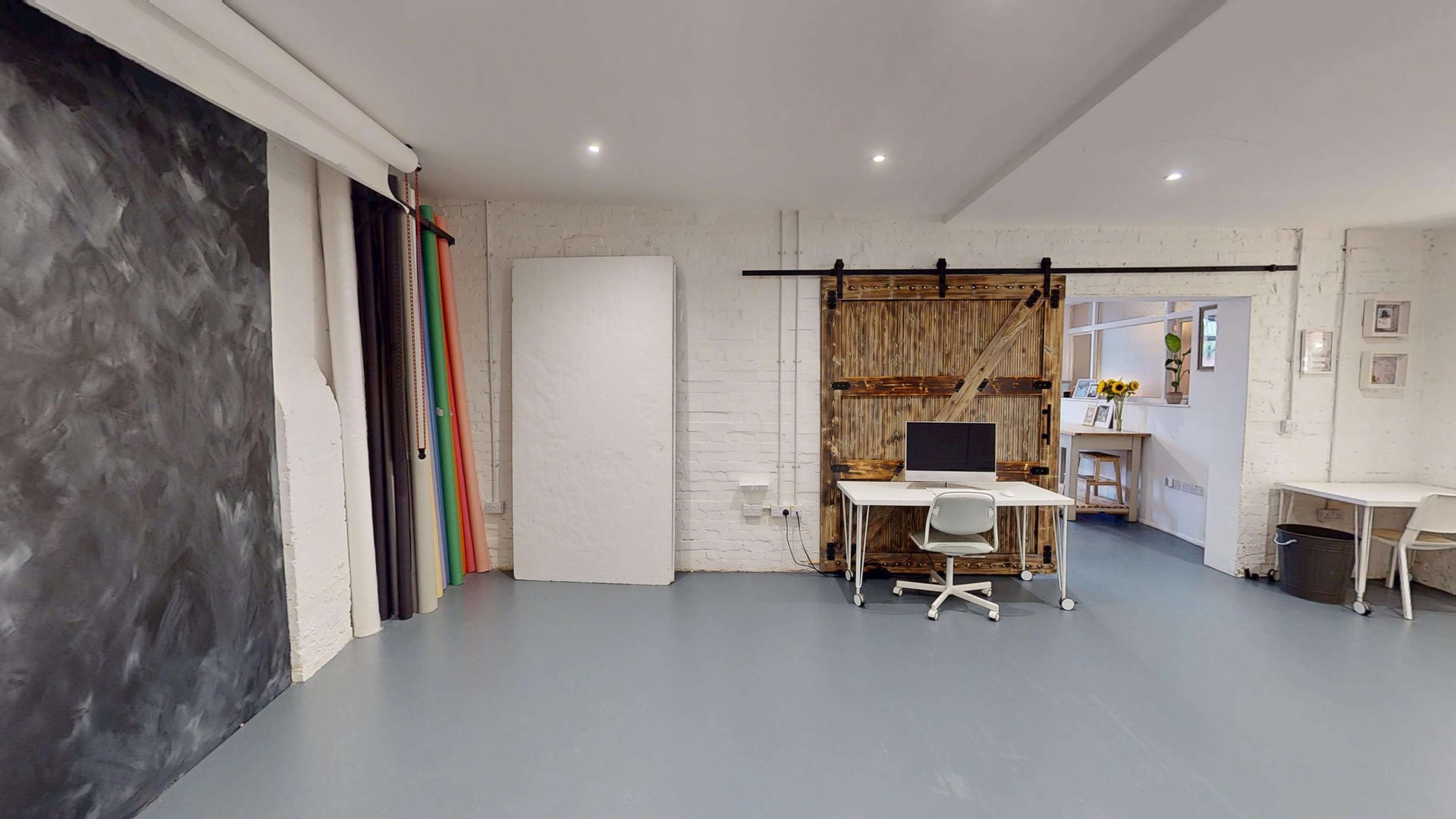 The image shows a creative workspace with a wooden desk and computer, surrounded by a white brick wall, a dark chalkboard, and colorful drapes.