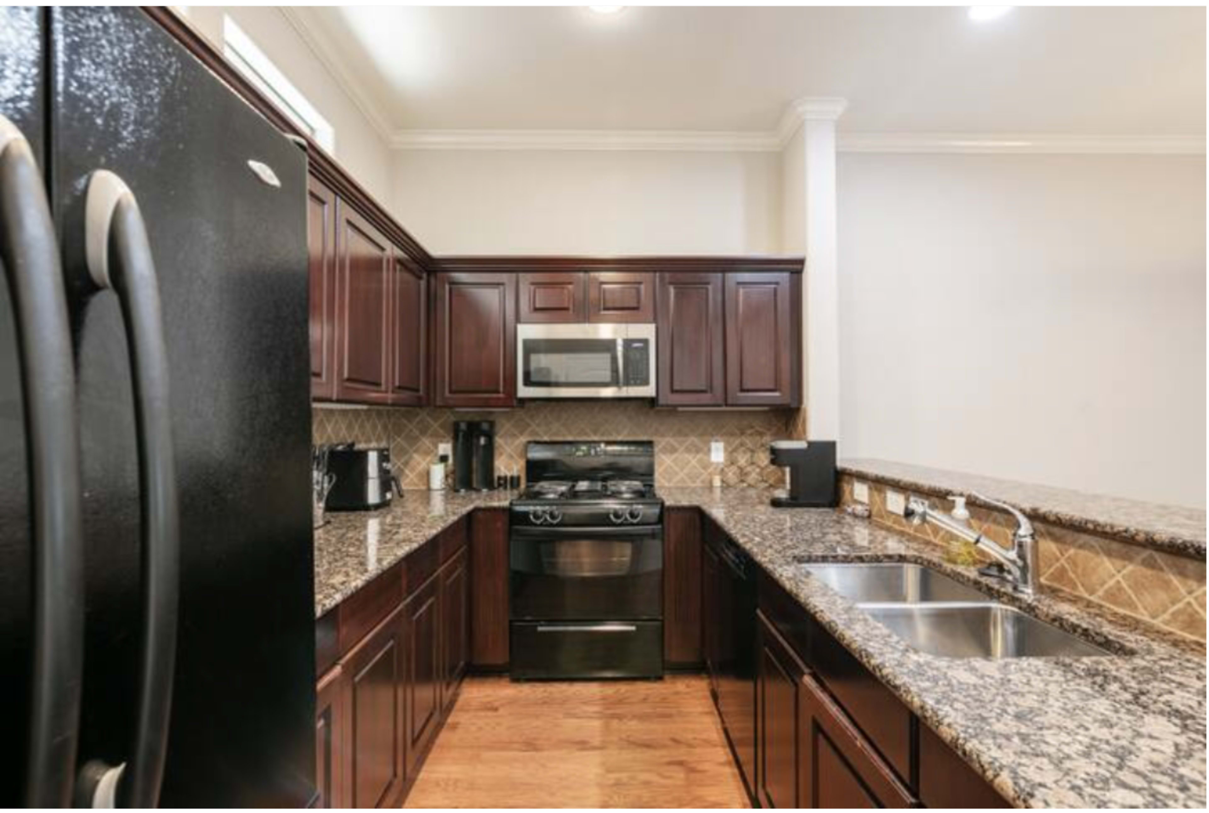 The image shows a modern kitchen with dark wooden cabinets, granite countertops, a stainless steel refrigerator, and a double sink.