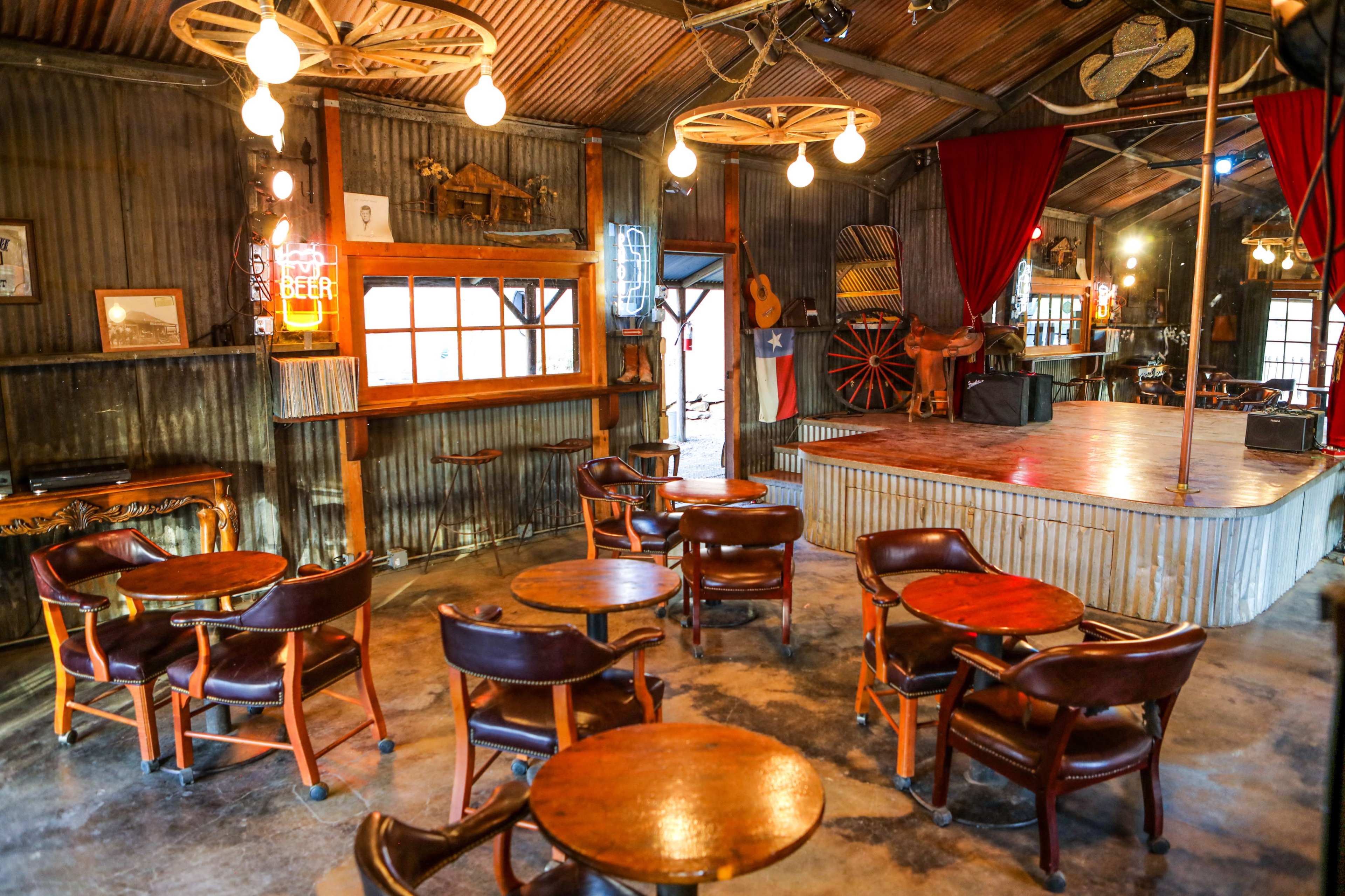 The interior of a rustic bar features wooden tables and chairs arranged around a central stage, with the walls adorned with various decorations and a beer sign illuminated in neon.