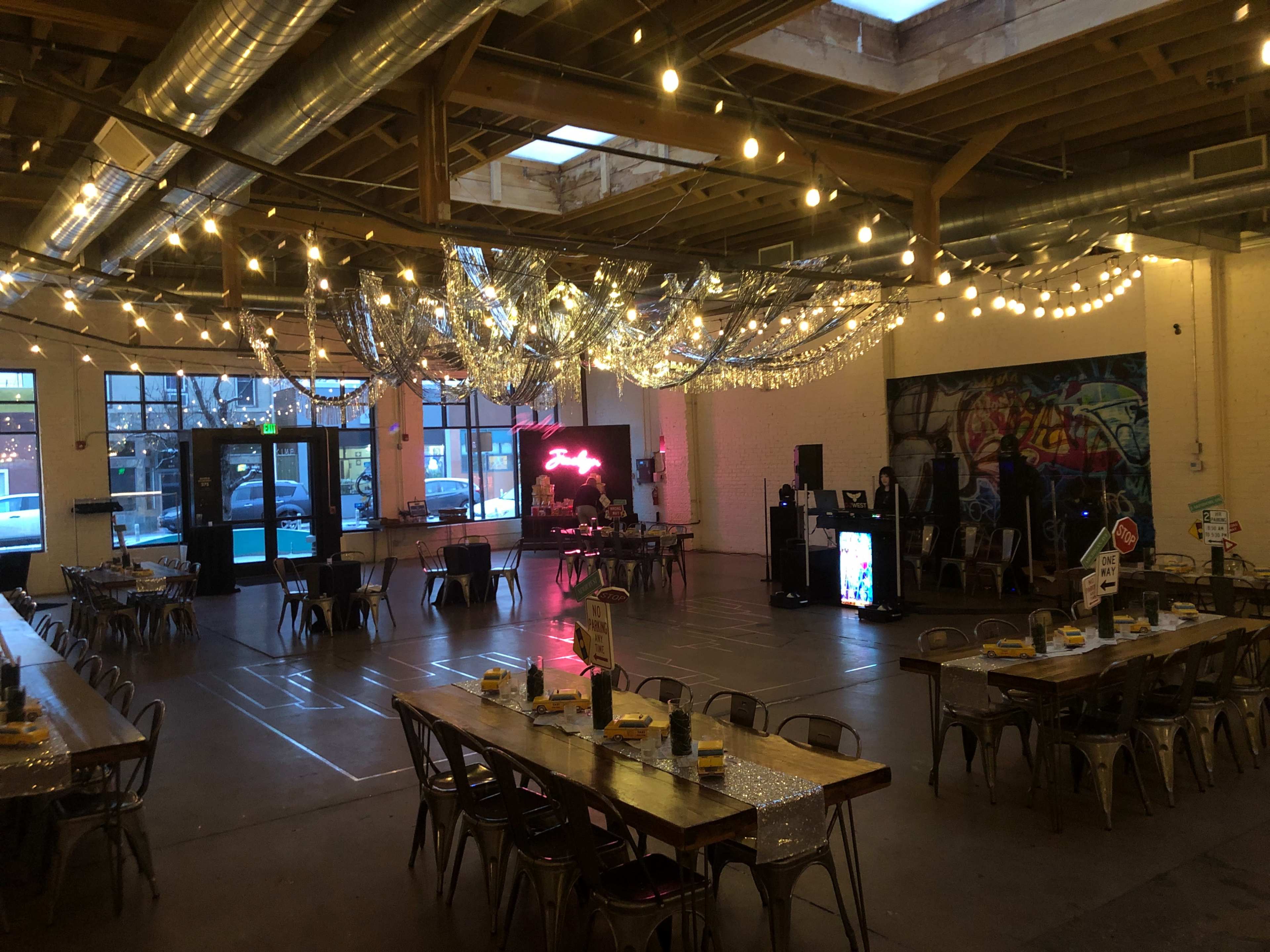 The image shows a spacious event venue with tables arranged in rows, string lights hanging from the ceiling, and a stage area featuring a neon sign.