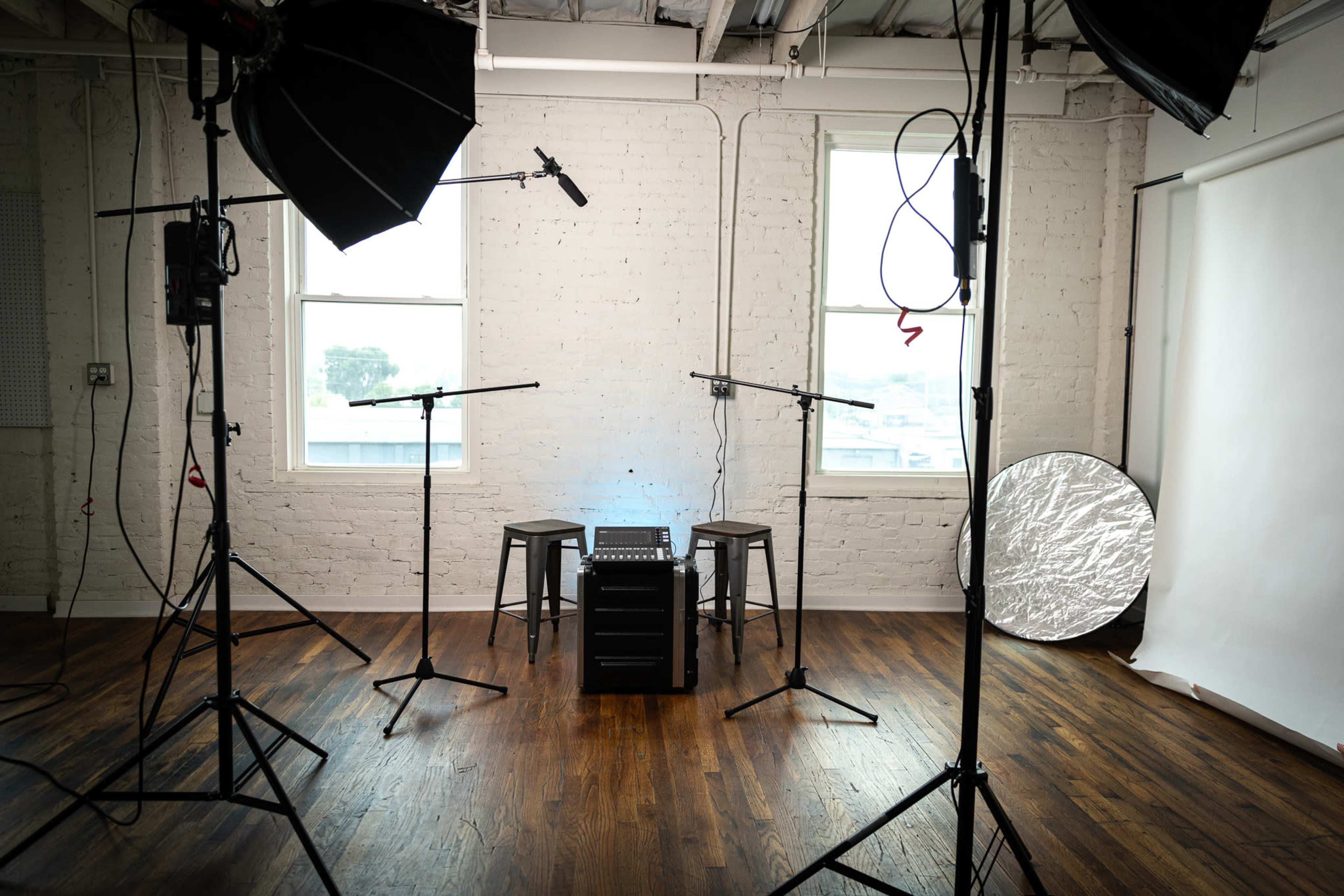 A photography studio is set up with two stools, a black equipment cart, microphones, and lighting gear positioned around a backdrop.