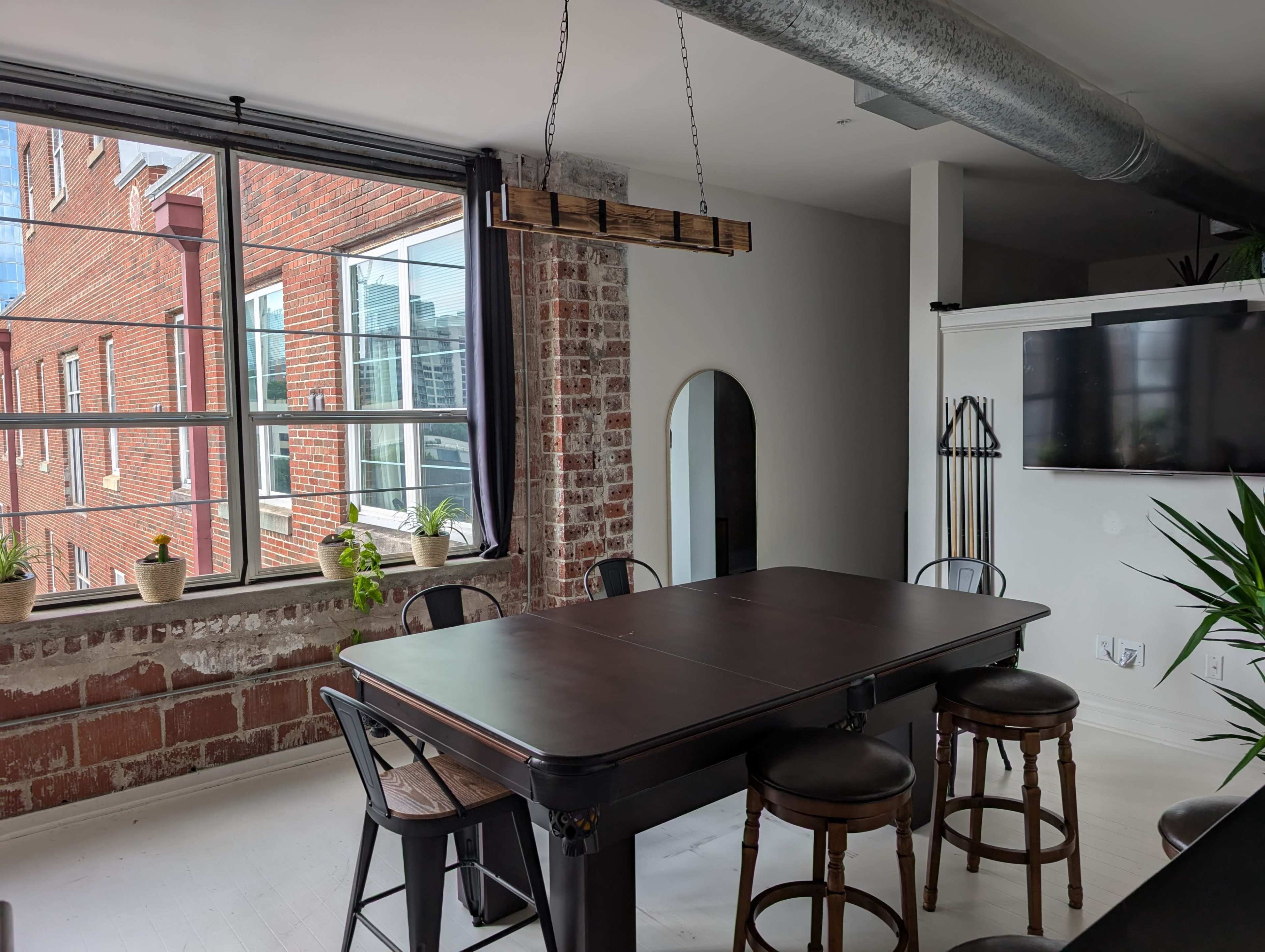 The image shows a dining area with a dark wooden table surrounded by black and wooden chairs, set against a backdrop of brick walls and large windows.