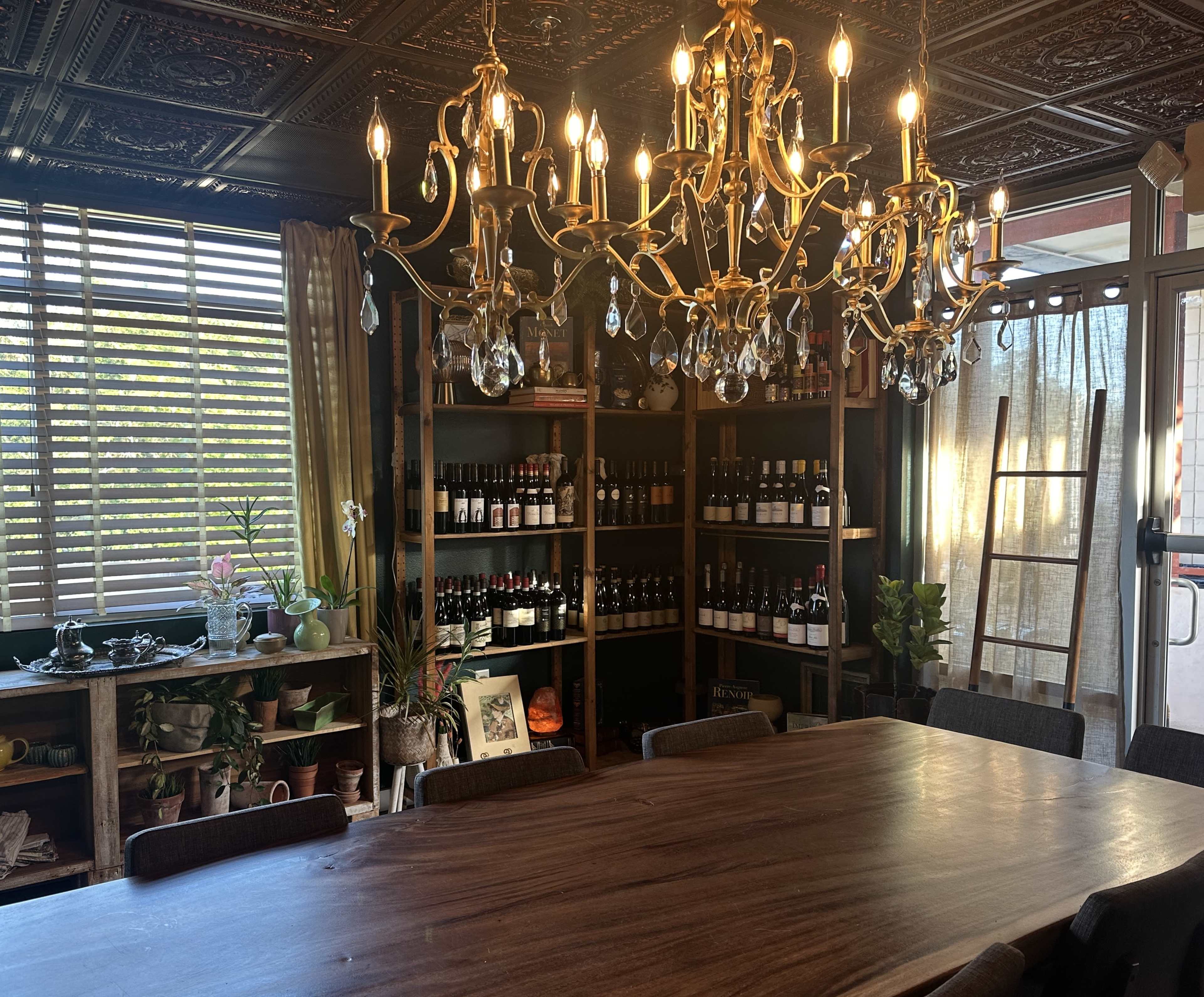 The image shows a dining area featuring a large wooden table, a chandelier with multiple lights, and shelves stocked with various wine bottles and decorative plants.