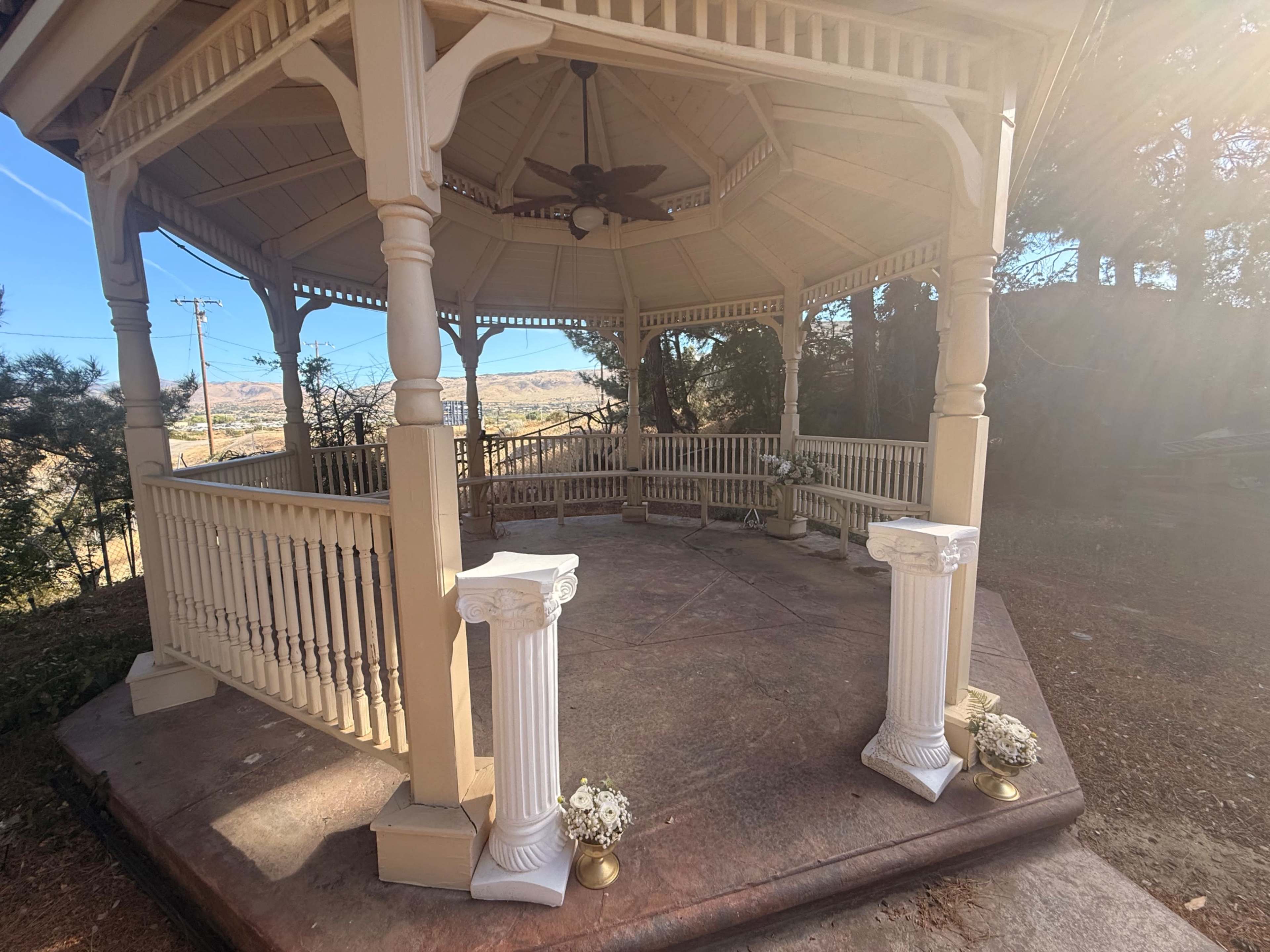 The image shows a wooden gazebo with a ceiling fan, situated on a stone platform surrounded by trees and open land in the background.