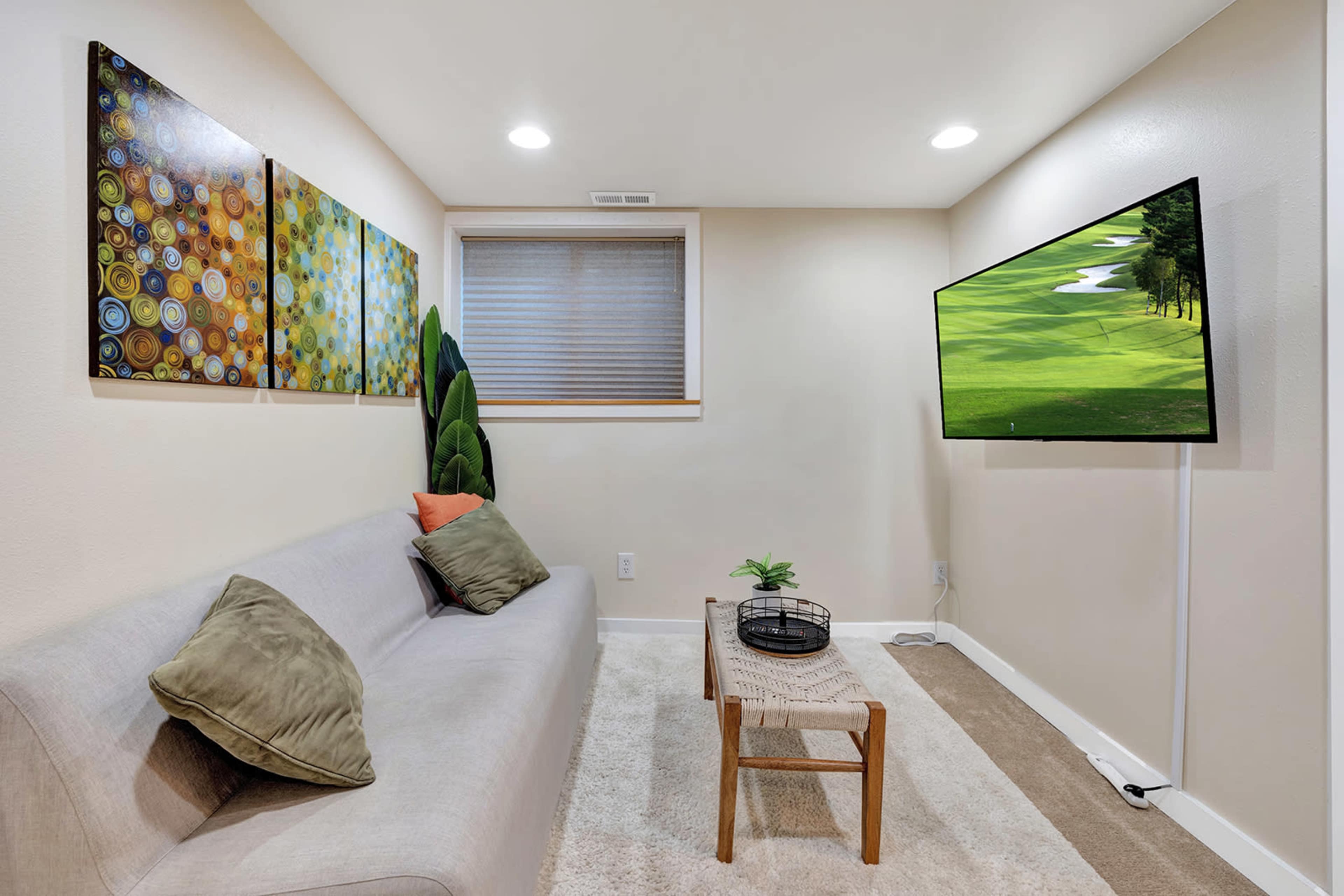 The image shows a cozy living space with a gray sofa, two abstract paintings on the wall, a flat-screen TV, and a small wooden table with decorative items.