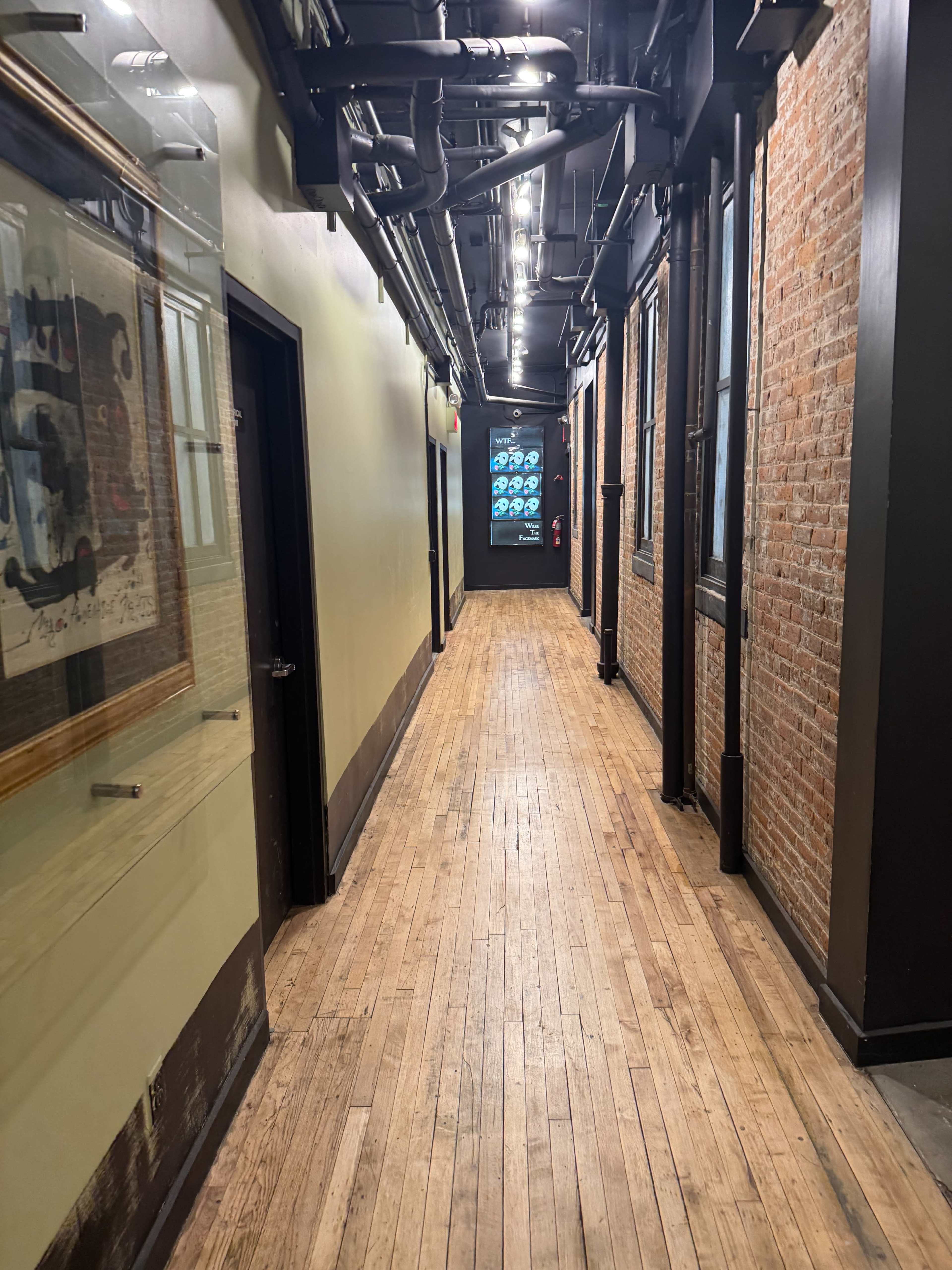 A long hallway features exposed brick walls, wooden flooring, and overhead pipes, with a glass-paneled door at the end.