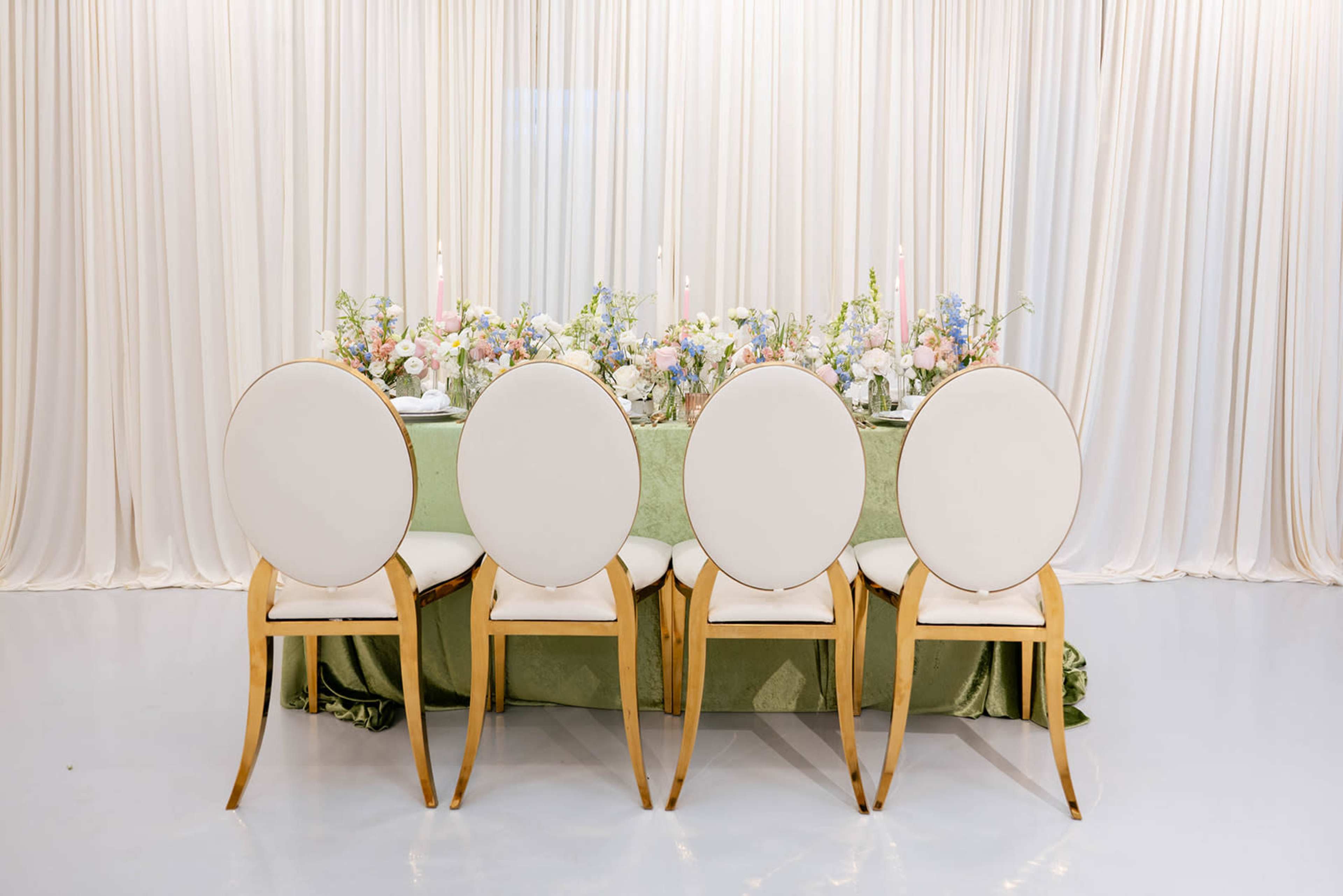 A set of four white chairs with round backs is arranged at a table decorated with floral arrangements and candles in front of a backdrop of cream-colored drapes.