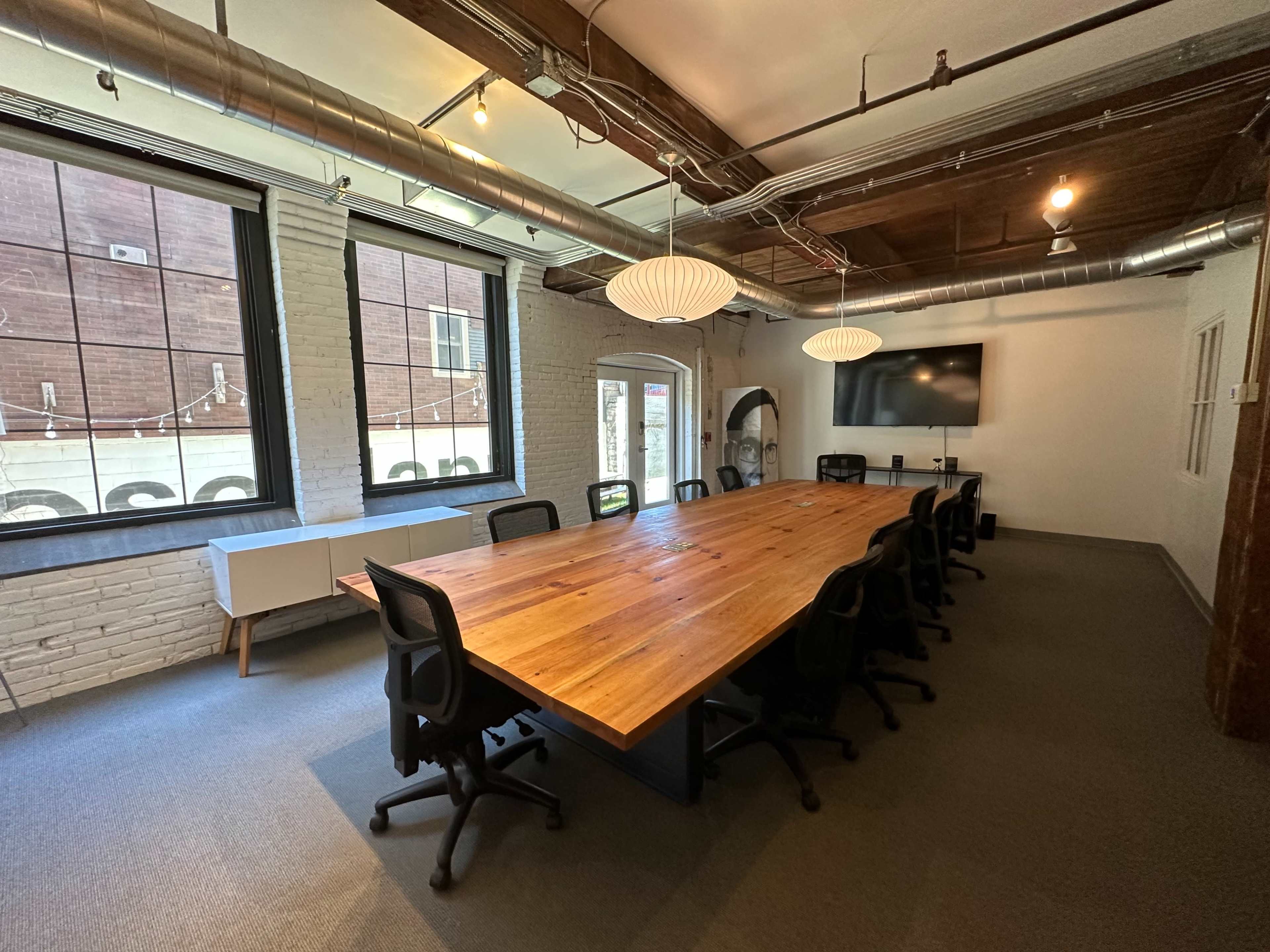 A large wooden conference table with black chairs is centered in a well-lit meeting room featuring exposed brick walls and multiple windows.