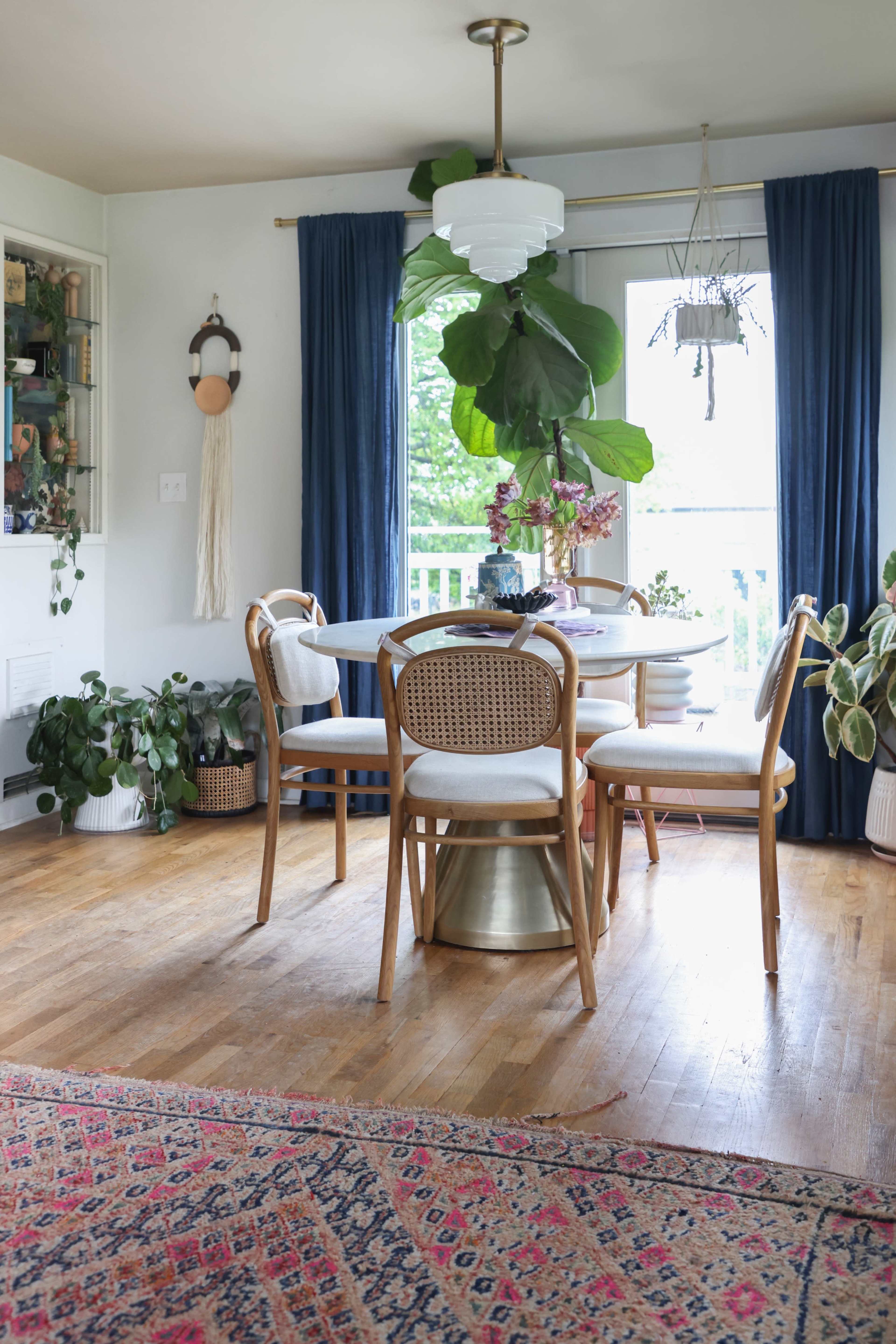 A dining area features a round table surrounded by four chairs, large leafy plants, and blue curtains, with a patterned rug on the wooden floor.