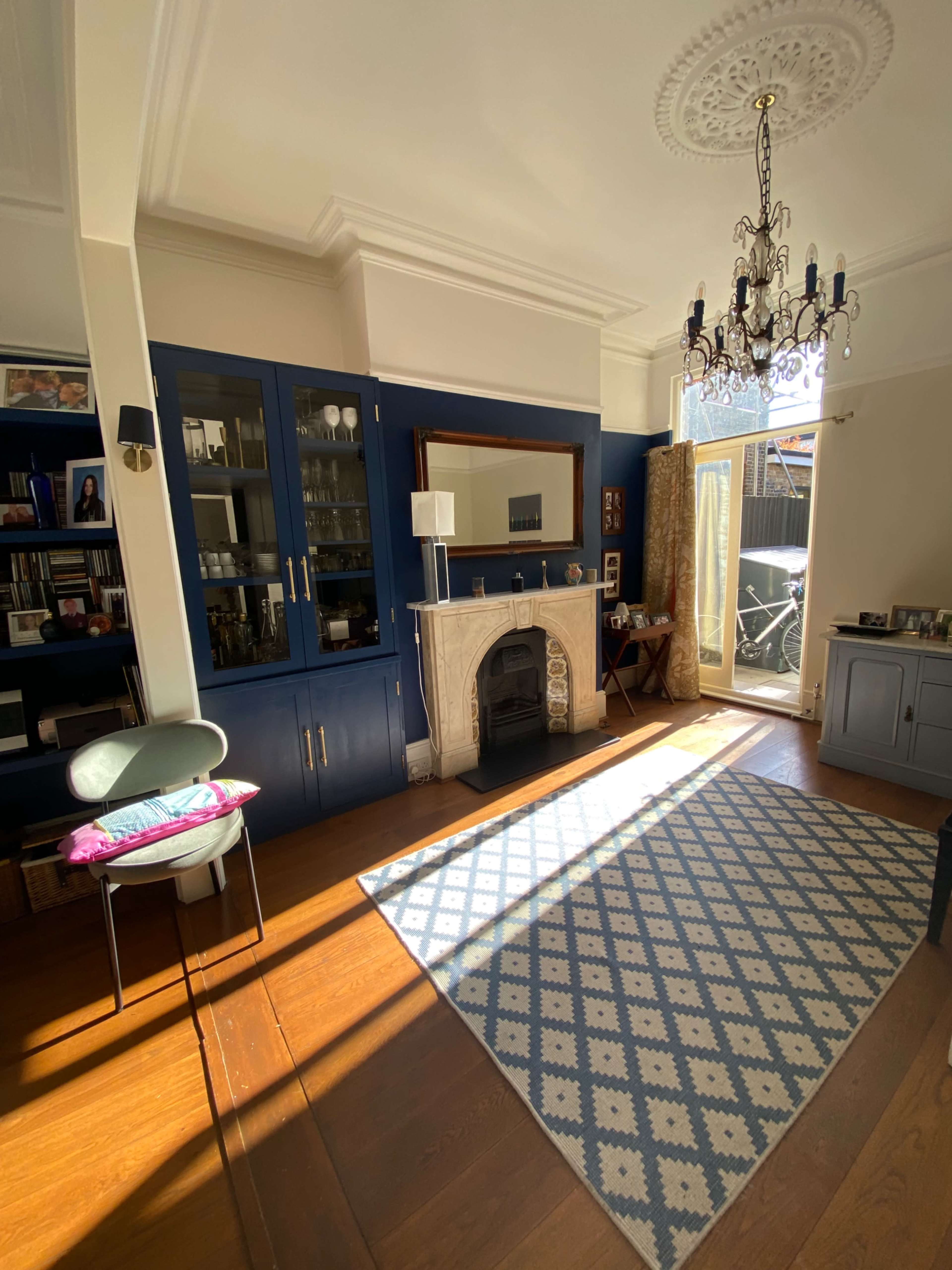 The image shows a well-lit living room with blue cabinetry, a decorative fireplace, and a patterned rug on the wooden floor.
