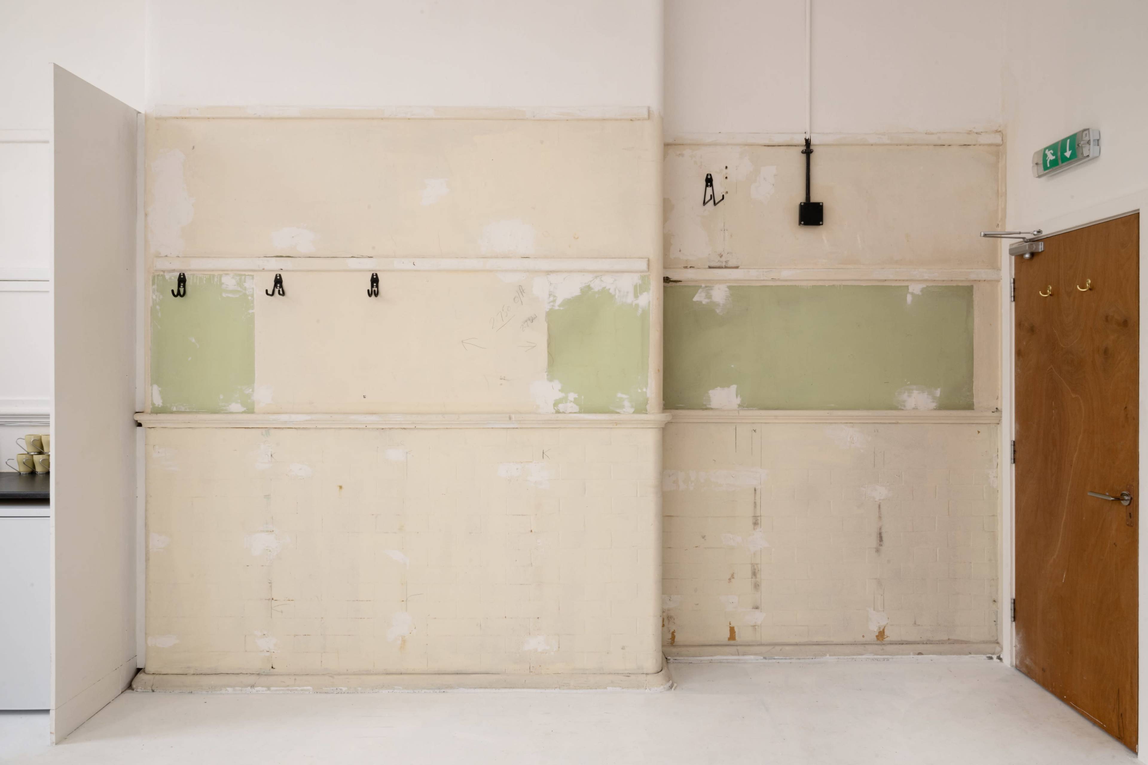 The image shows an unfinished interior wall with peeling paint, exposed sections, and several wall hooks.