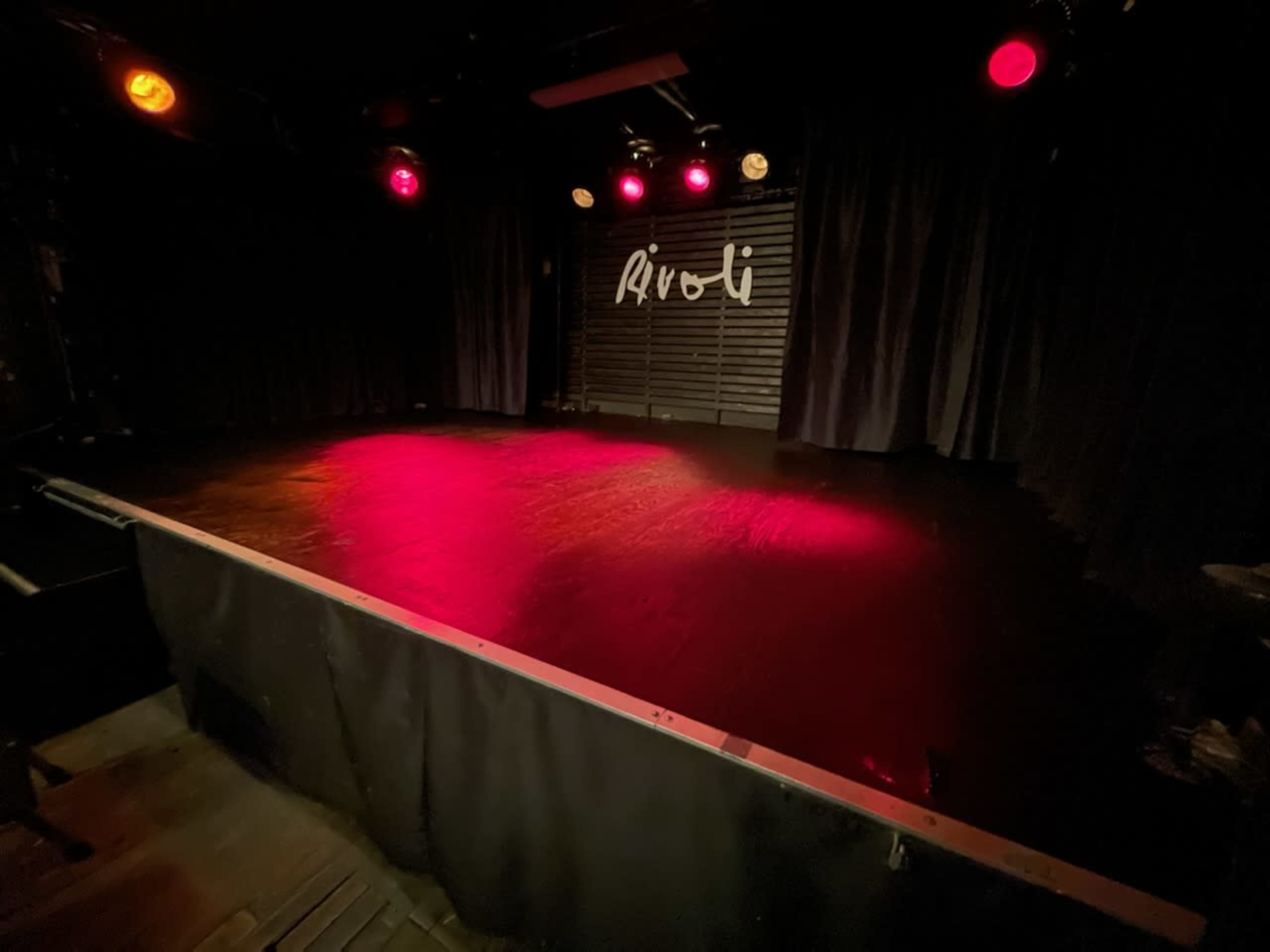 The image shows a dimly lit stage with red and white lights, featuring a backdrop that says "Rivoli."