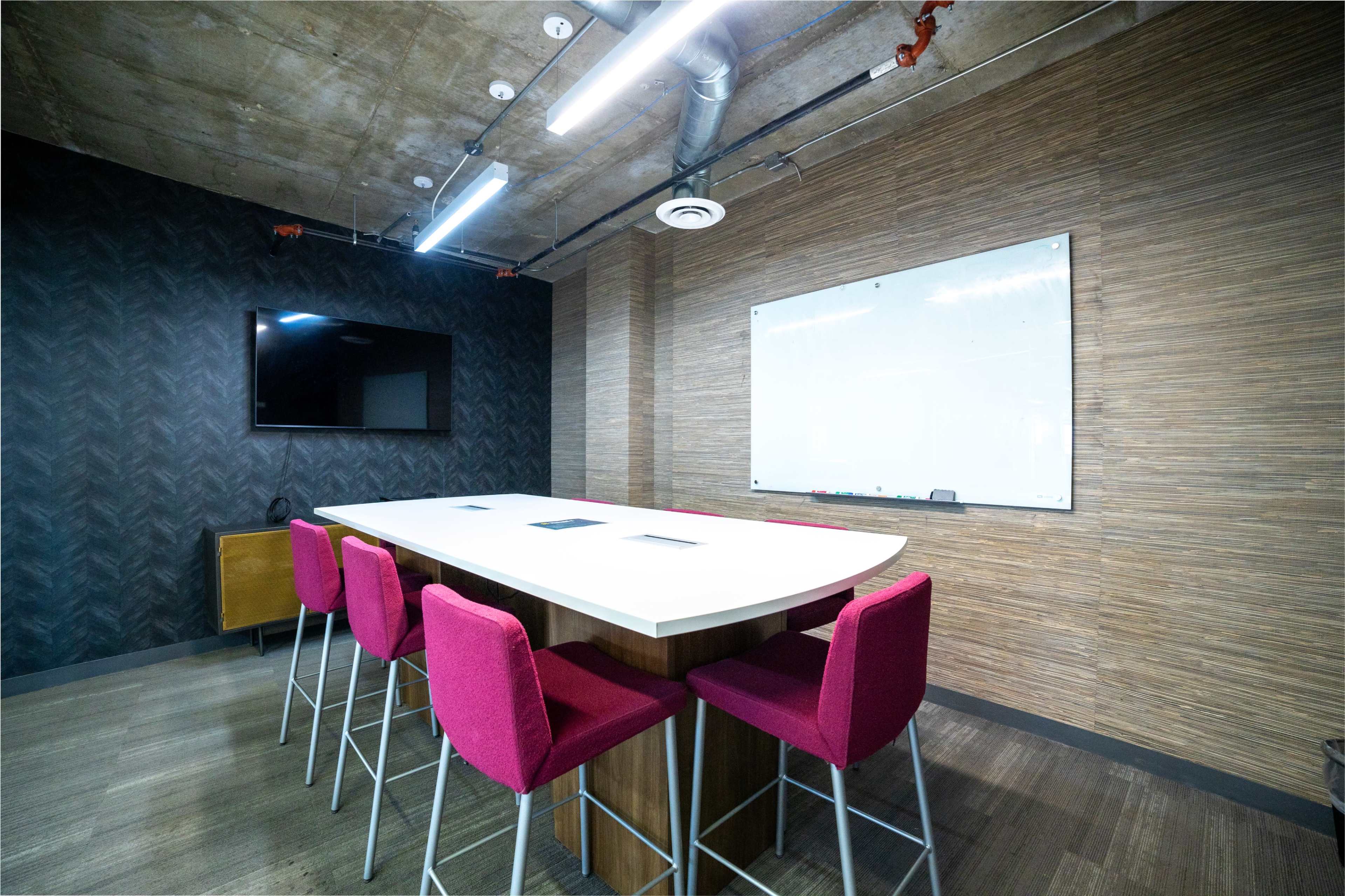 A modern meeting room features a long, white table surrounded by pink bar stools, a flat-screen TV, and a whiteboard on a textured wall.