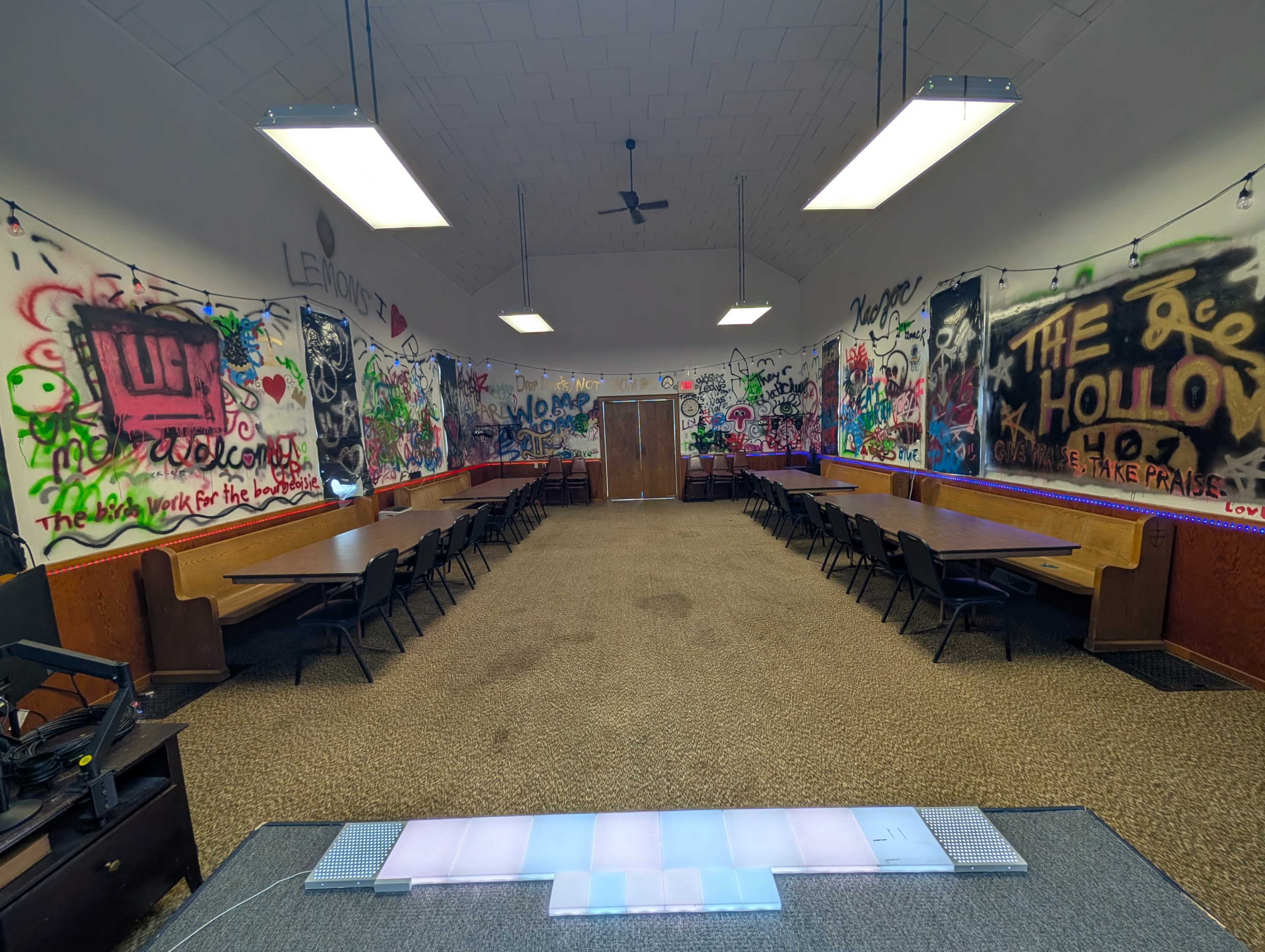 A large room with multiple long tables lined up against the walls, covered in colorful graffiti and equipped with overhead lighting.