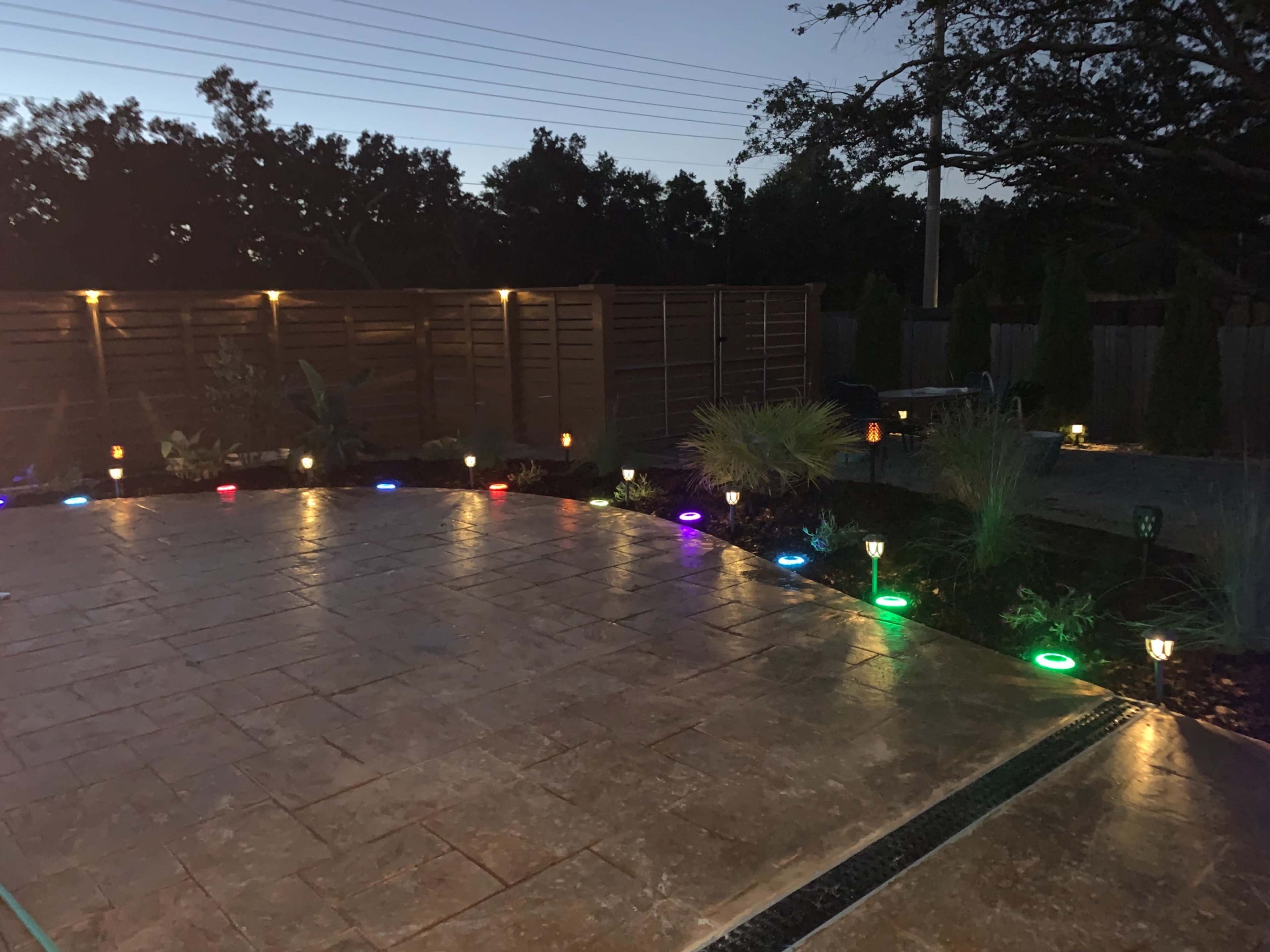 A patio area illuminated by colorful lights along the edges, surrounded by plants and a wooden fence.
