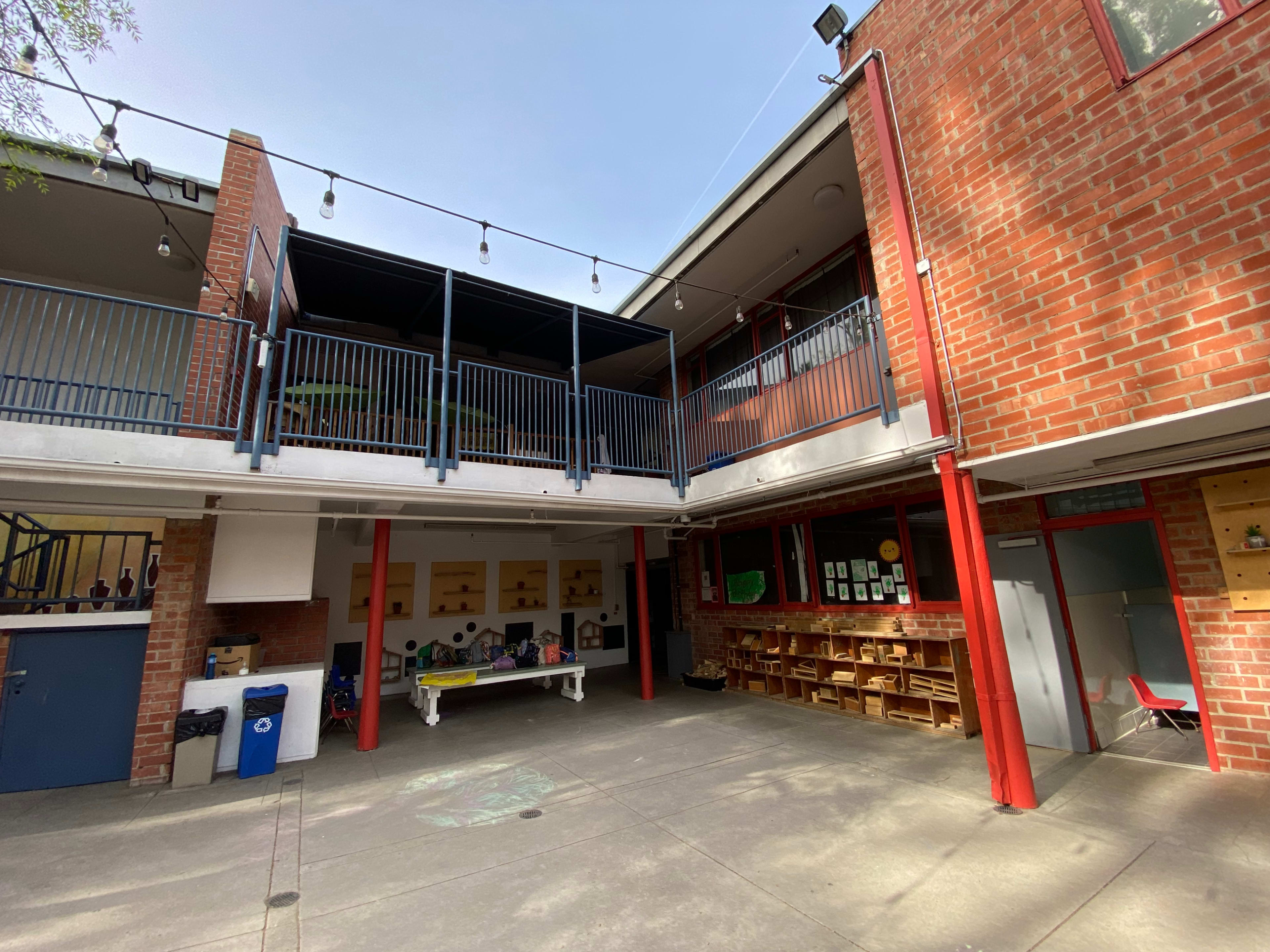 The image shows a two-level school building with red brick walls, featuring a balcony and a courtyard area filled with tables and art supplies.