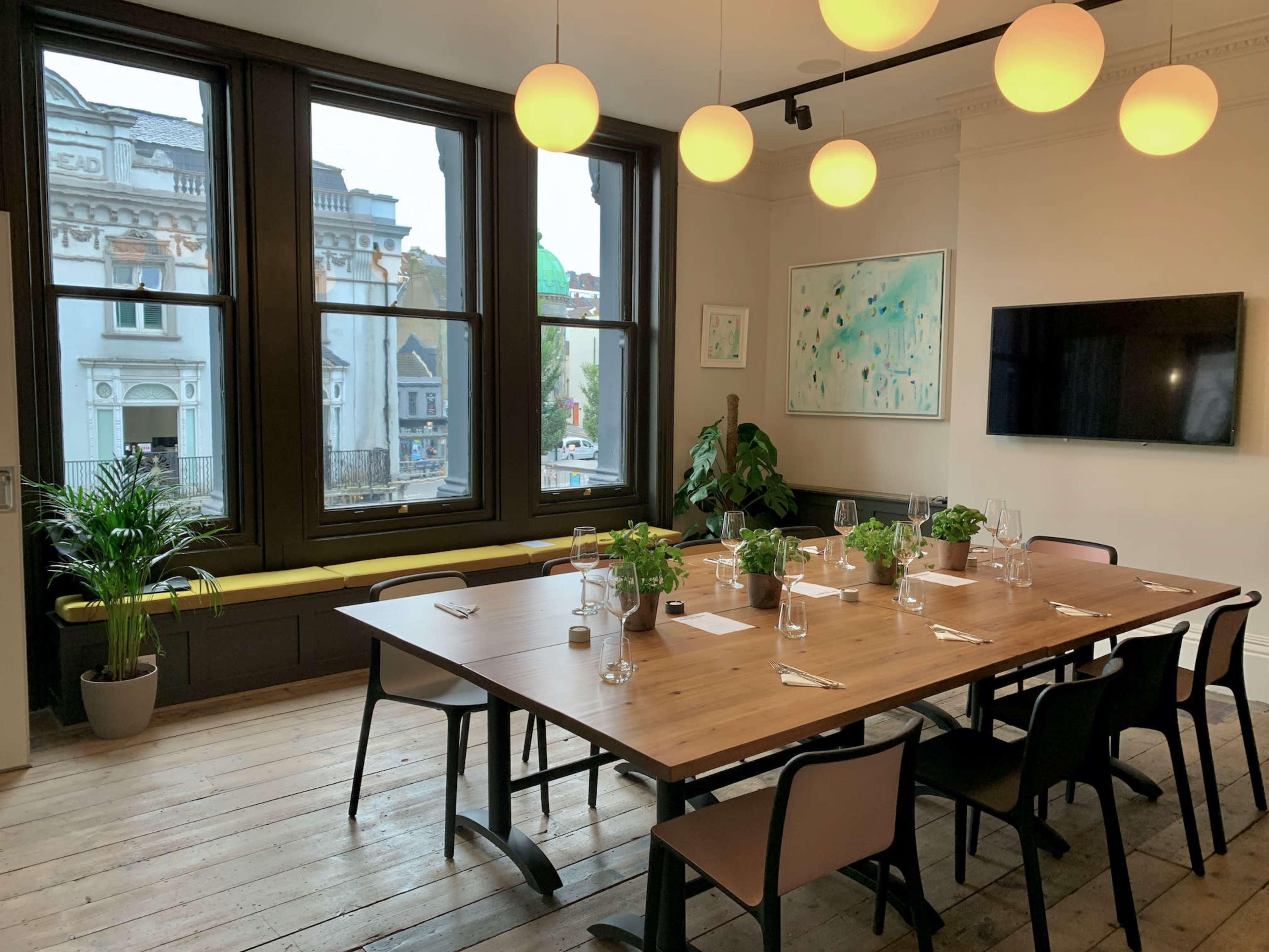 A dining room features a large wooden table set for a meal, surrounded by chairs, with large windows revealing an outdoor view and green plants in the corners.