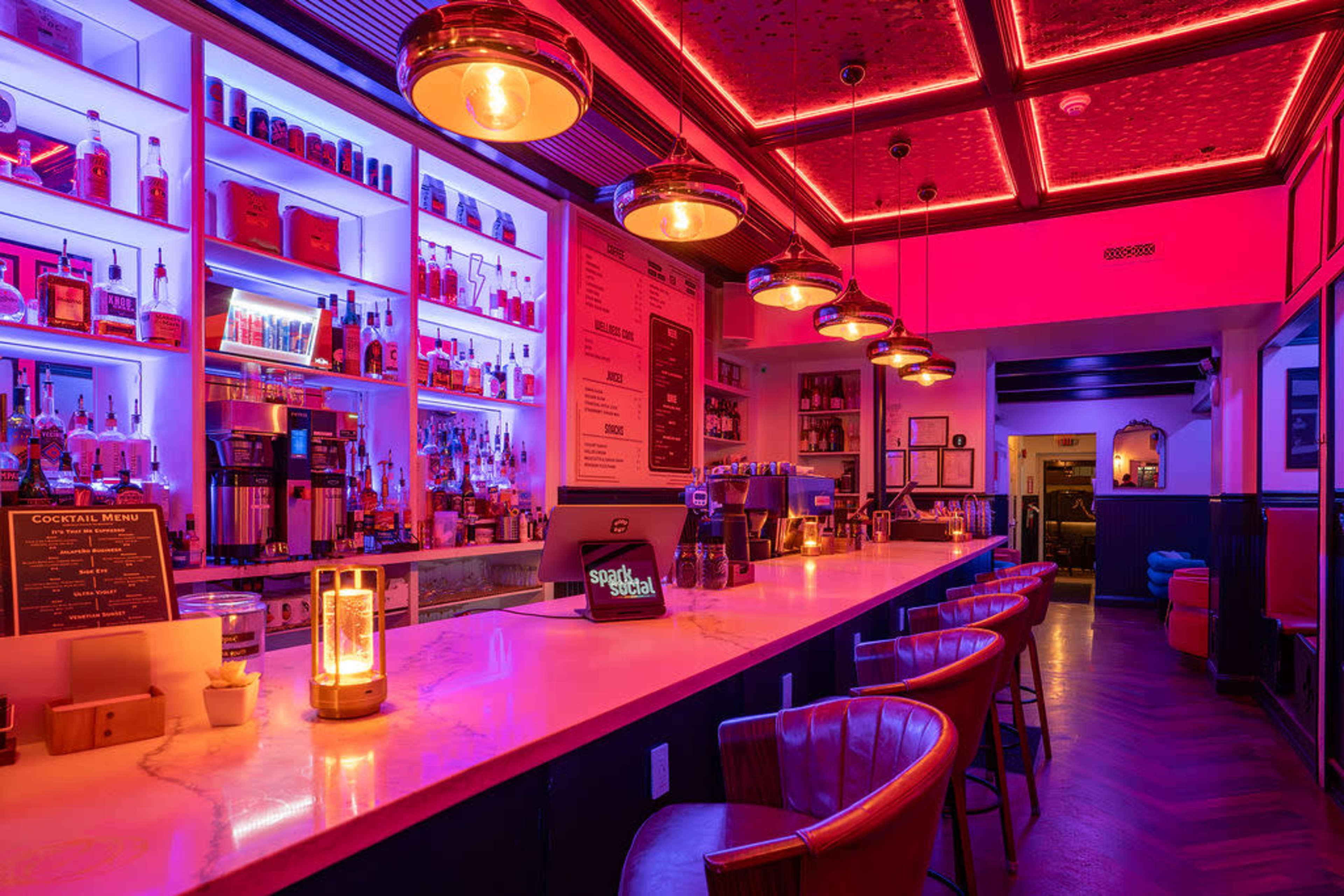 The image shows a stylish bar with illuminated shelves displaying various bottles and a marble countertop, accented by vibrant pink and blue lighting.