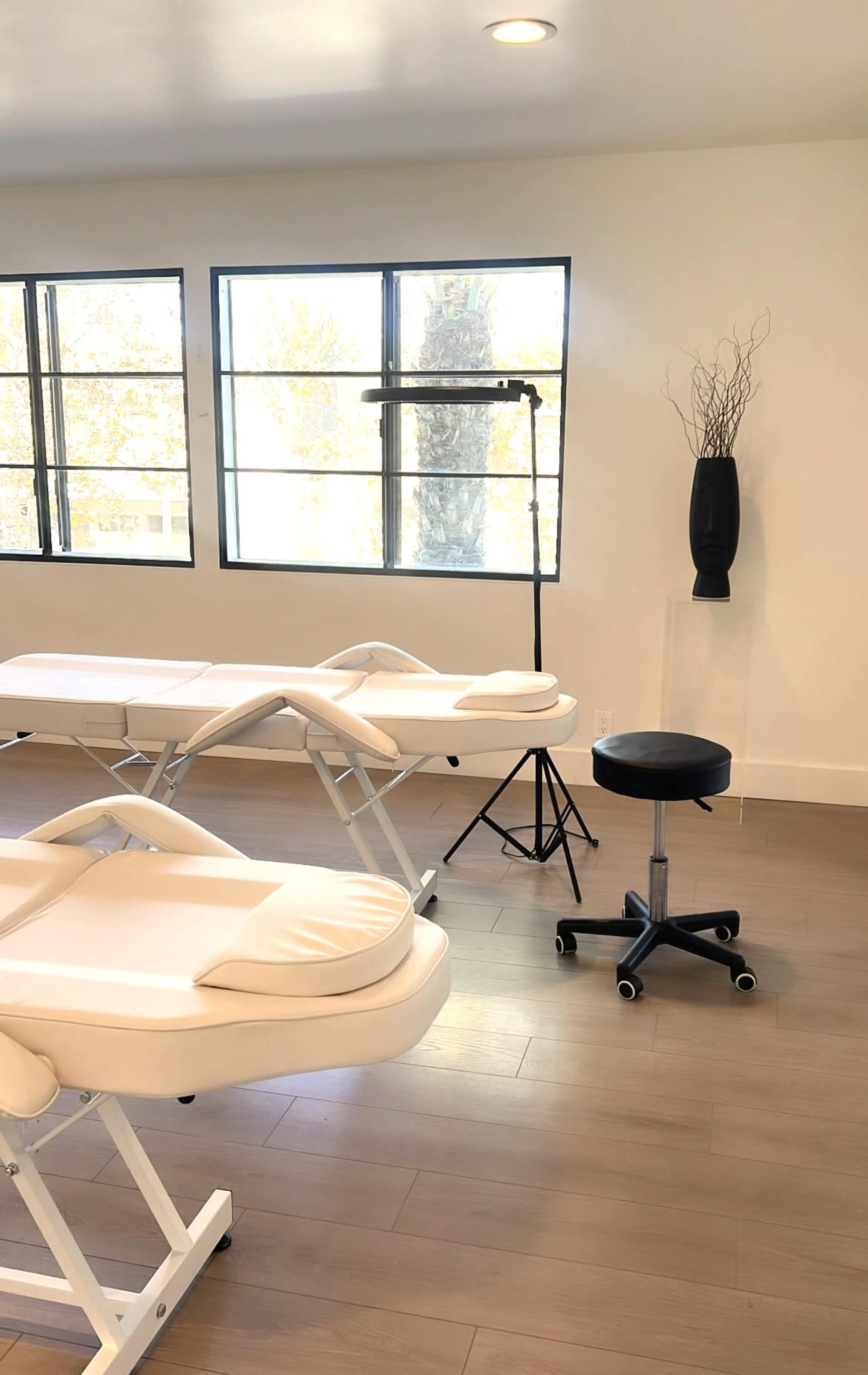A bright room with two massage tables, a stool, and a decorative vase near large windows.