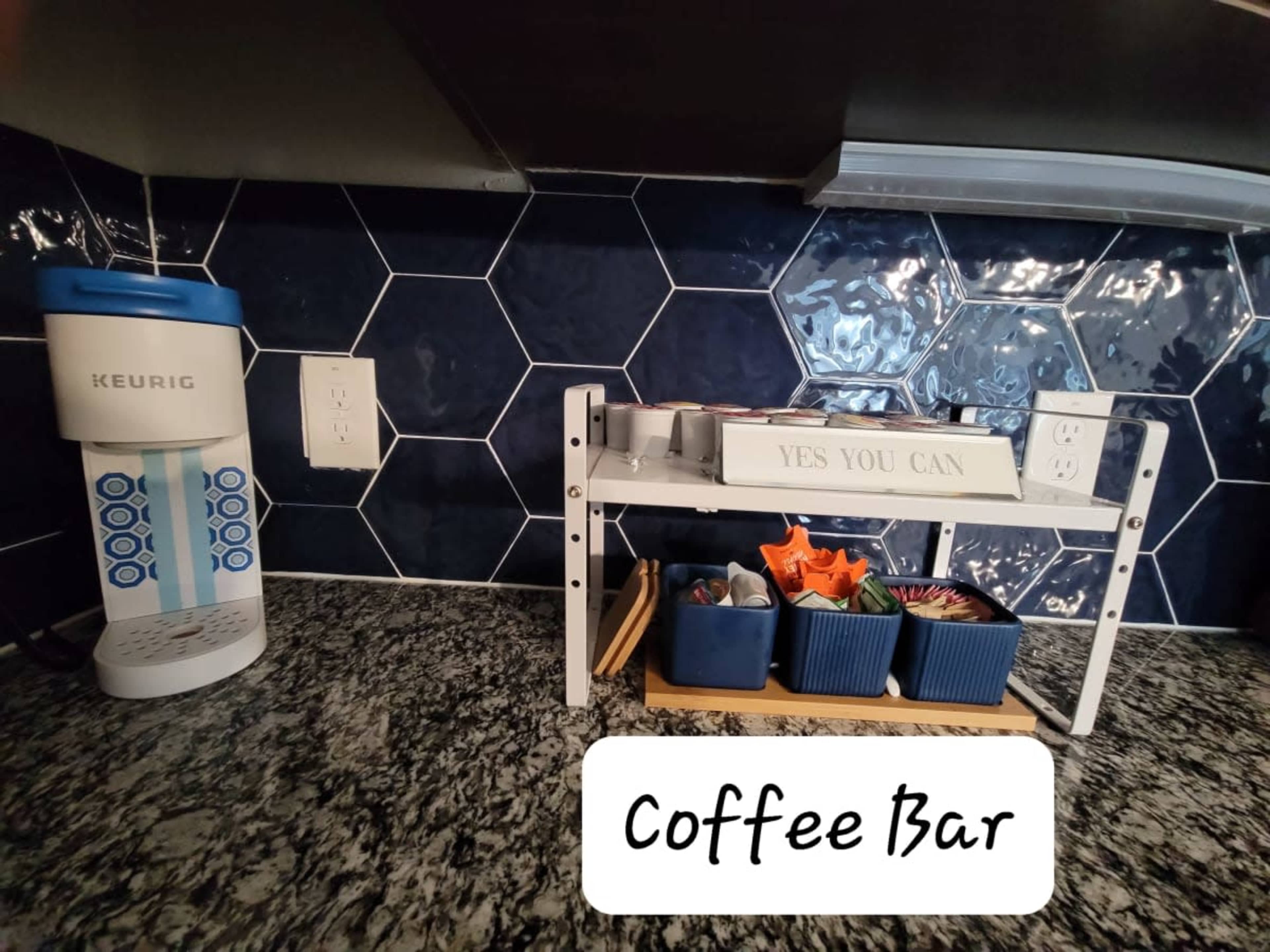A coffee bar setup featuring a Keurig coffee maker on the left and a tiered organizer with small containers on the right, all placed against a hexagonal tiled backsplash.