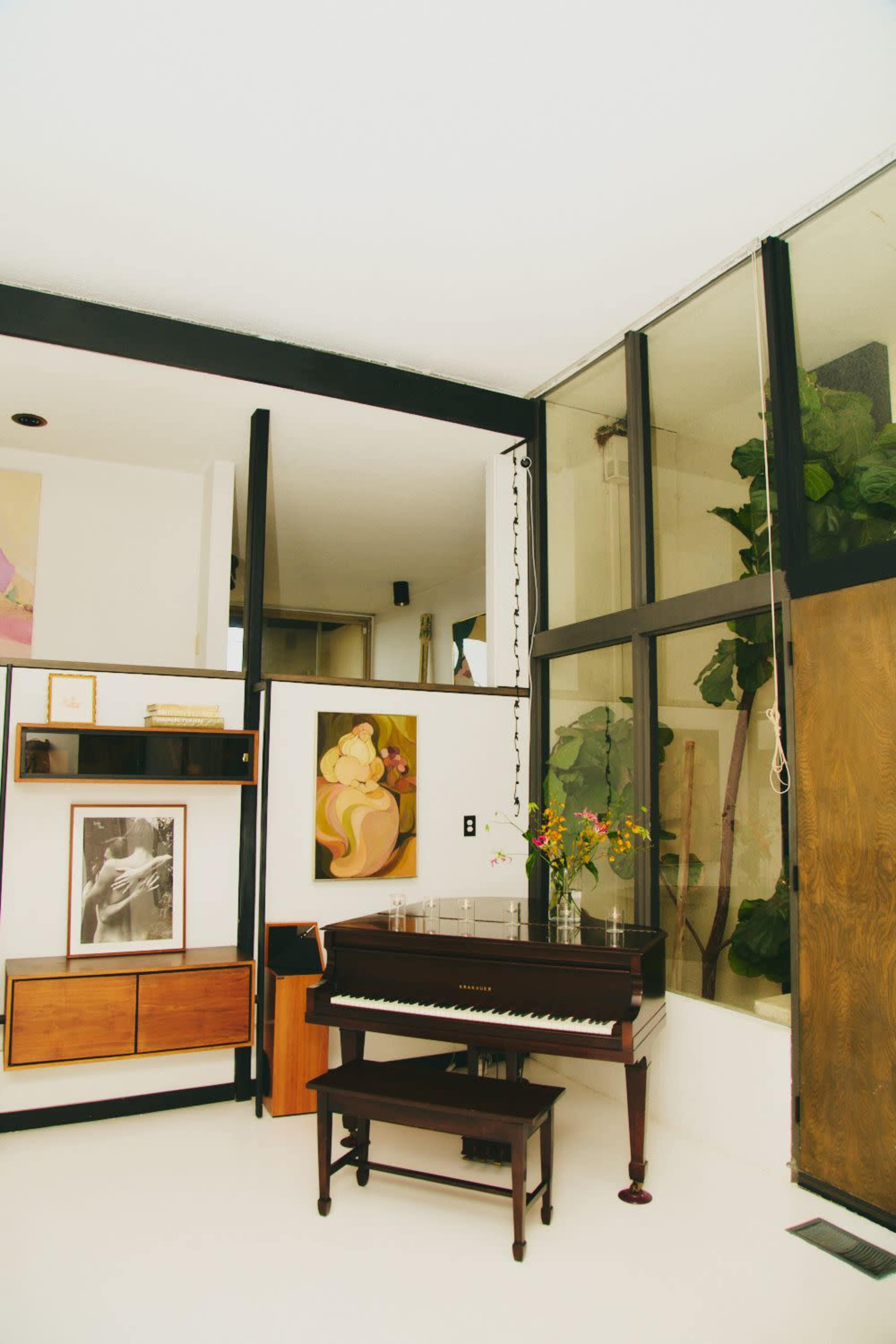 A grand piano is positioned in a sunlit room with large windows and artwork displayed on the walls.