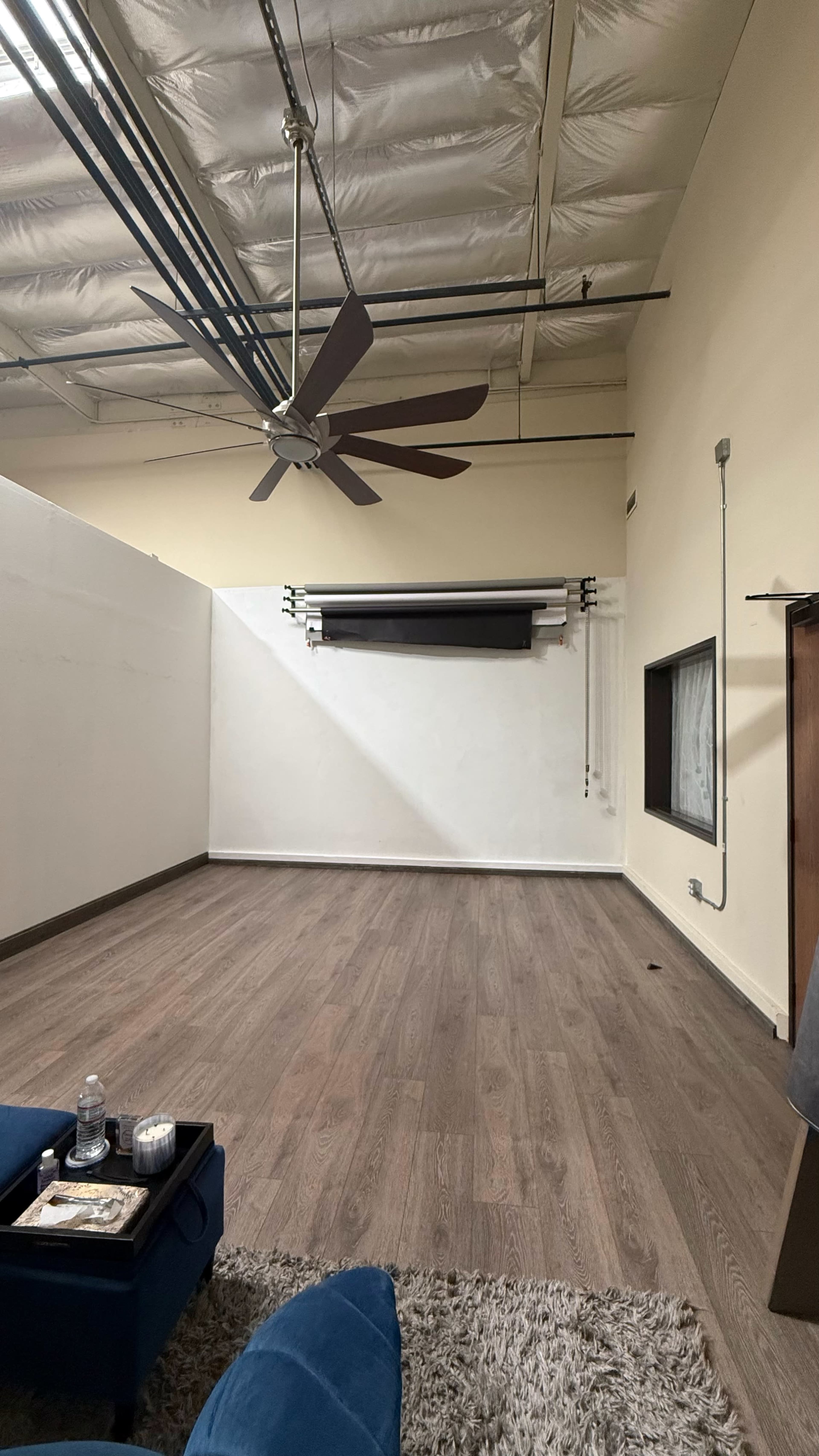 The image shows a spacious, empty room with a ceiling fan, a large window, and a backdrop setup against one wall.