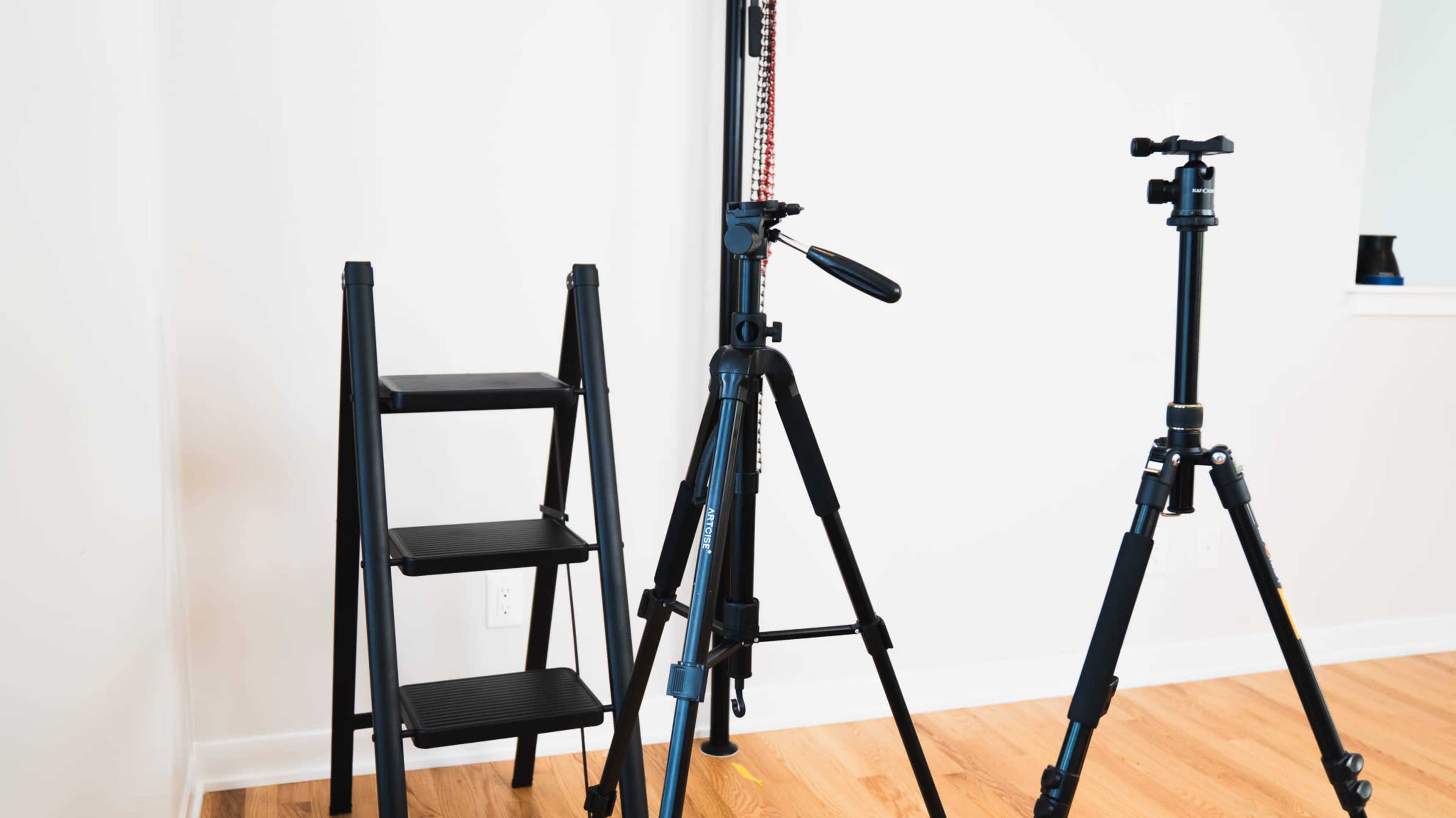 A room with a ladder, a photography tripod, and a second tripod positioned on a wooden floor.