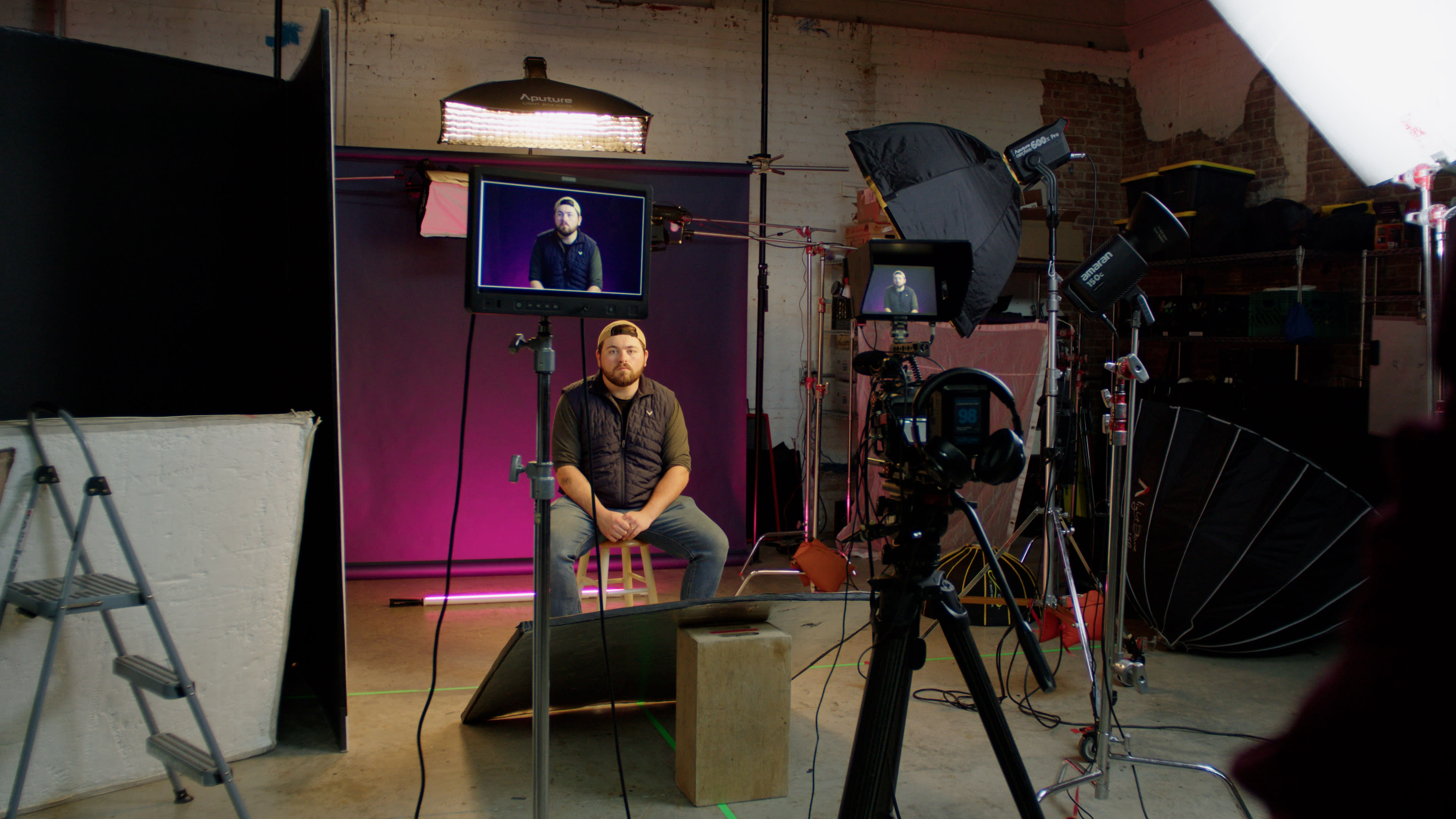A man is seated on a stool in a photography studio, surrounded by various lighting and camera equipment.