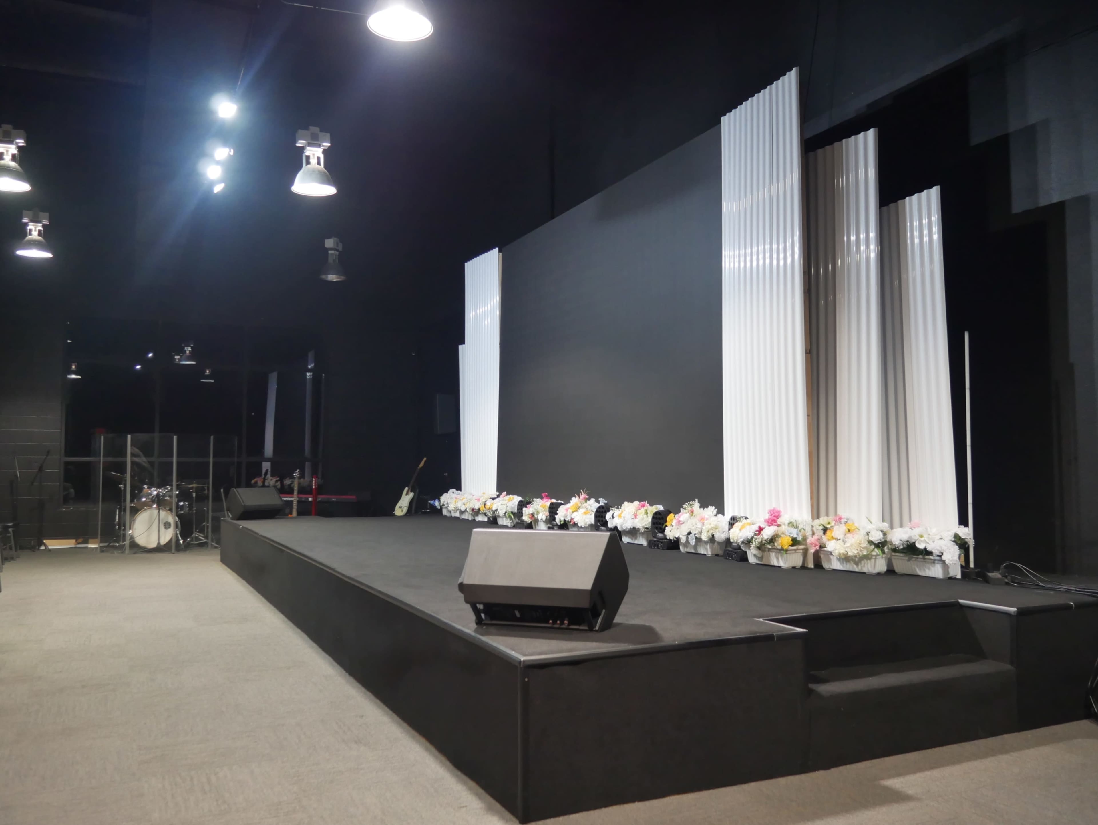 The image shows a stage set up with a black backdrop, white drapes, and flower arrangements along the front, in a darkened room designed for events.