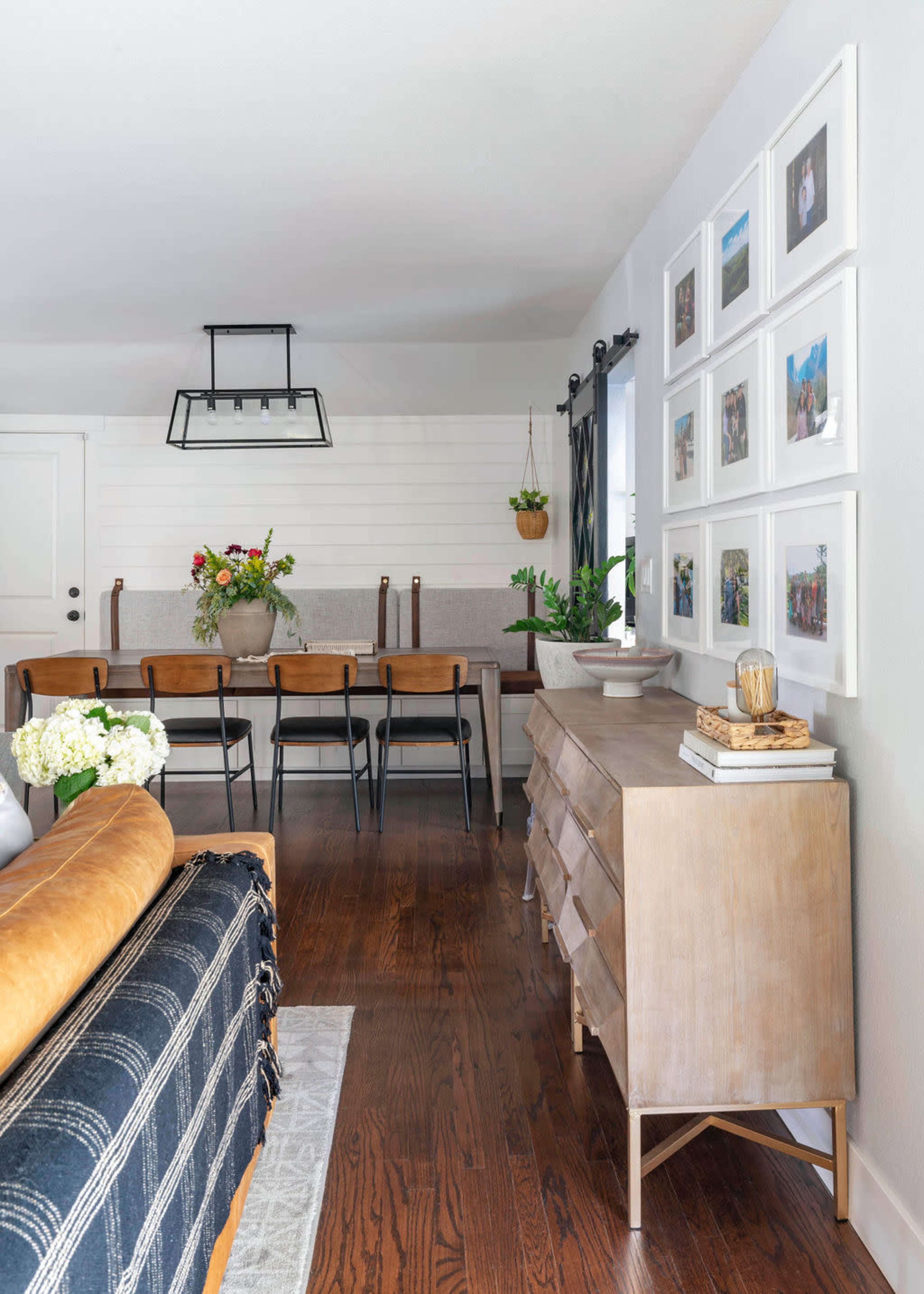 A living space featuring a leather sofa, a wooden sideboard, a dining table with black chairs, and framed photographs on the wall.