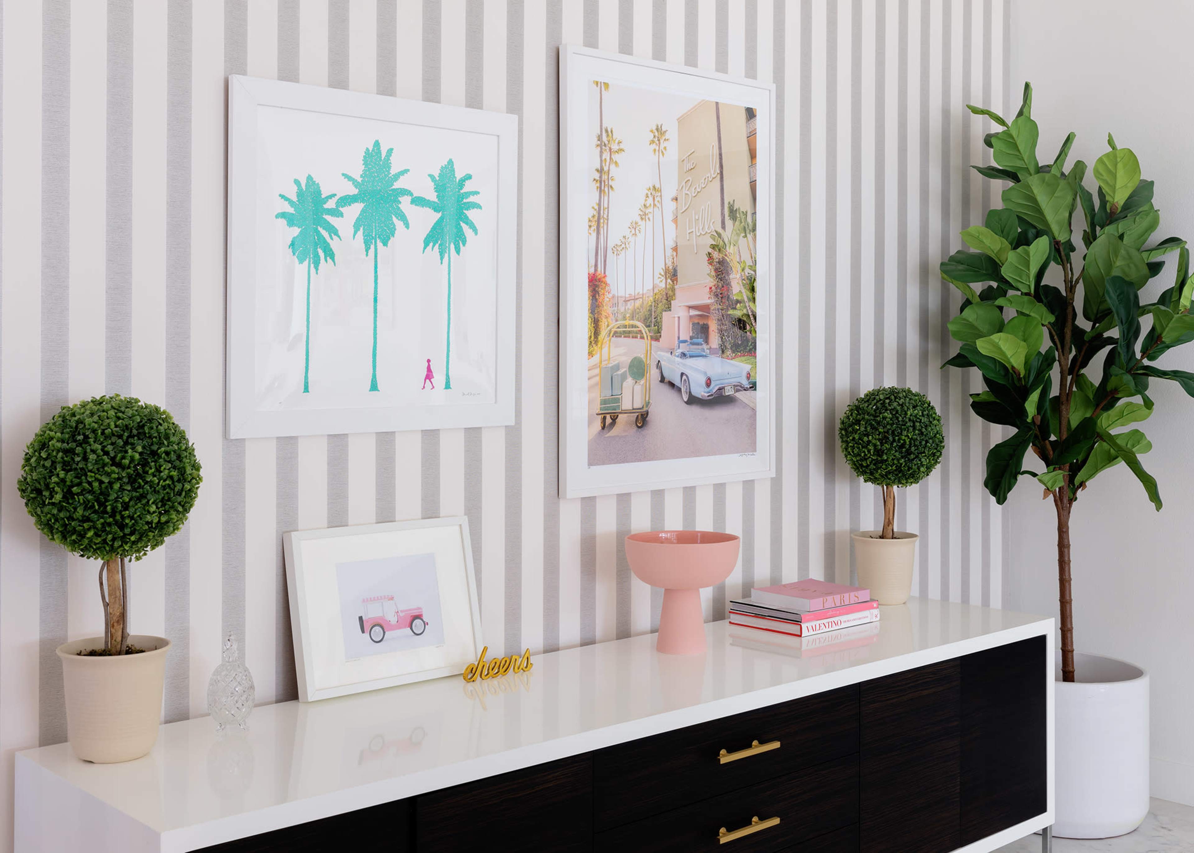 A modern sideboard displays framed artwork of palm trees and a pastel scene, accompanied by decorative plants and various decorative items.