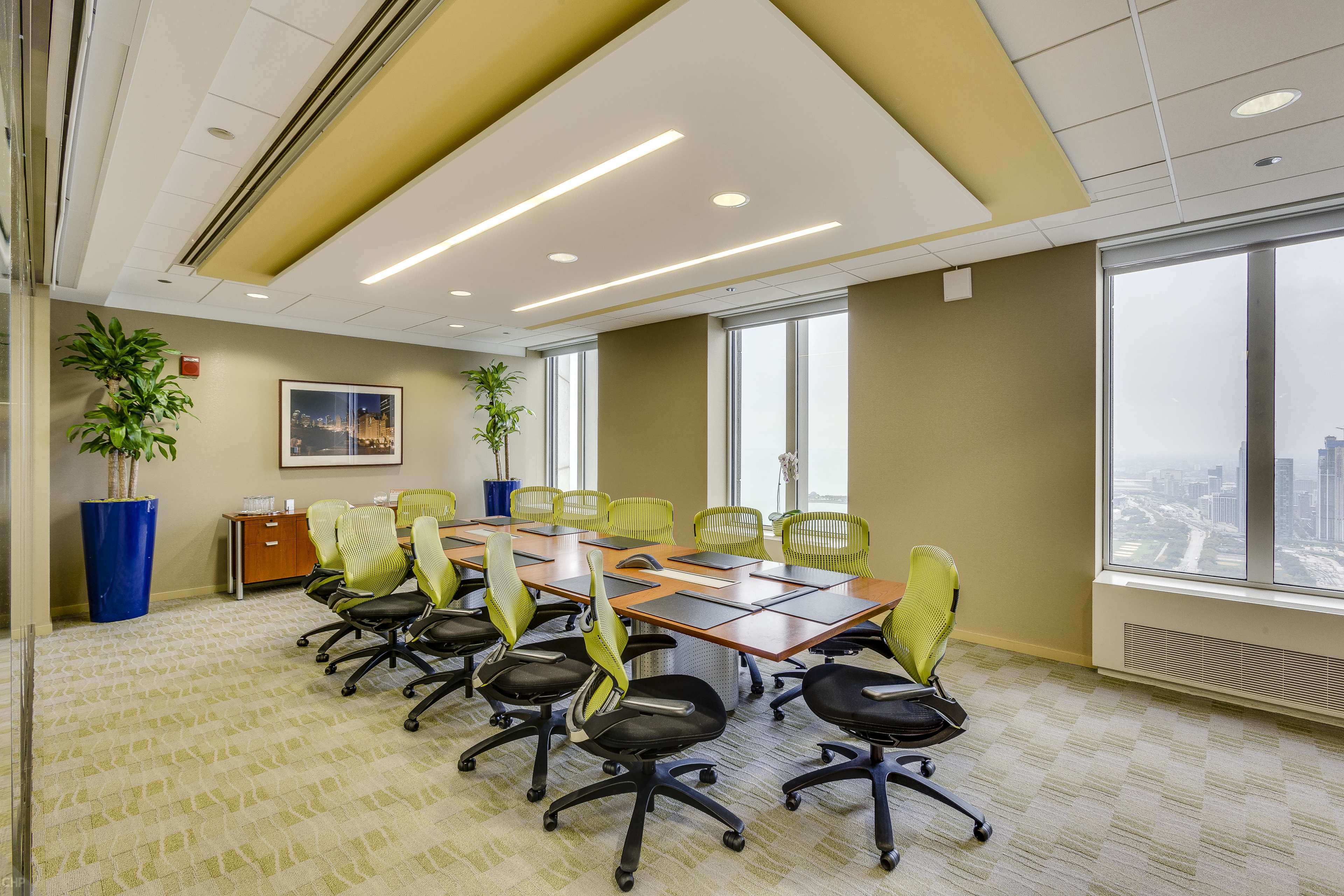 A modern conference room features a long table surrounded by green office chairs, with large windows showing a city view and potted plants in the corners.