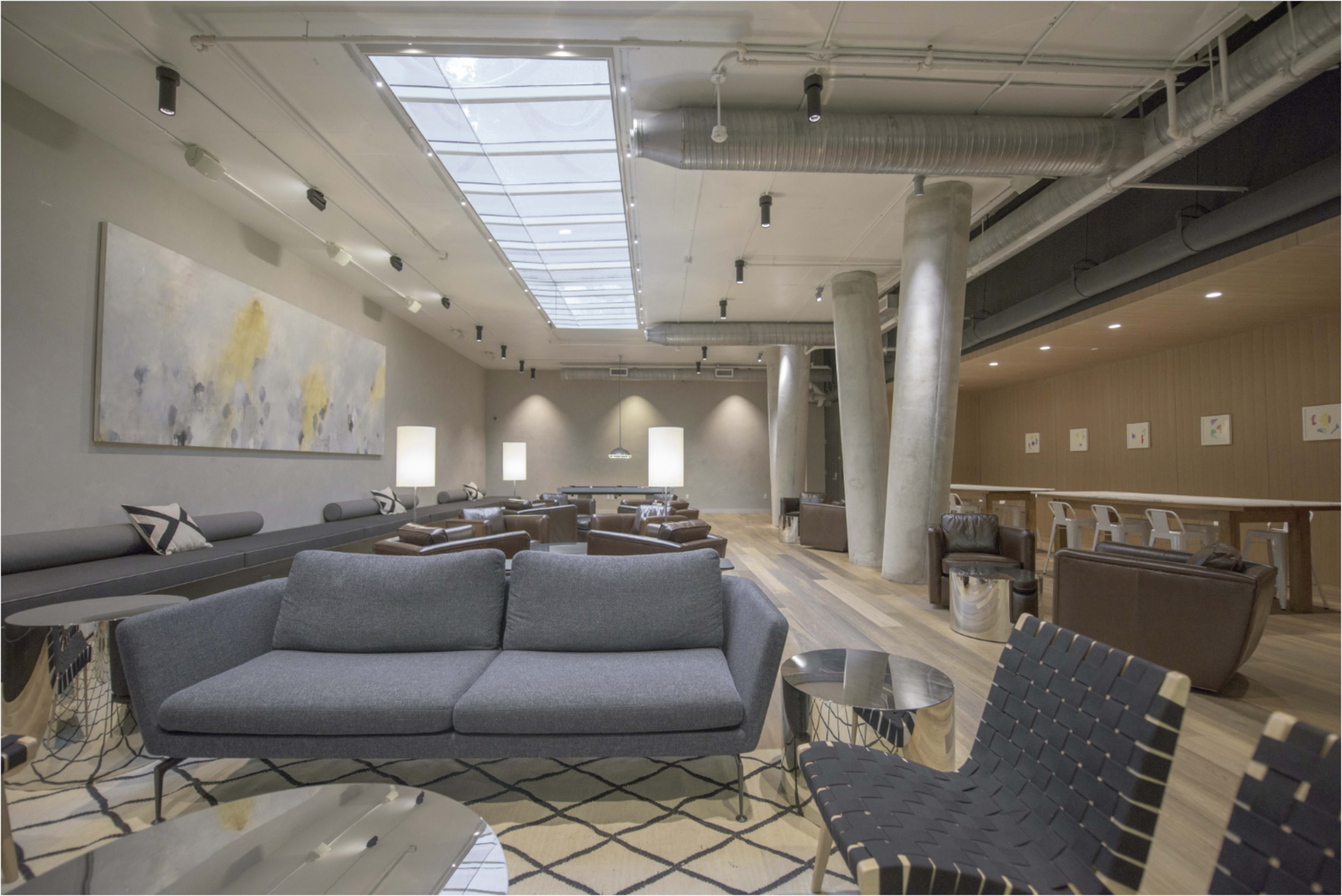The image shows a spacious modern lounge area with gray sofas, wooden flooring, and a bar in the background under skylights.