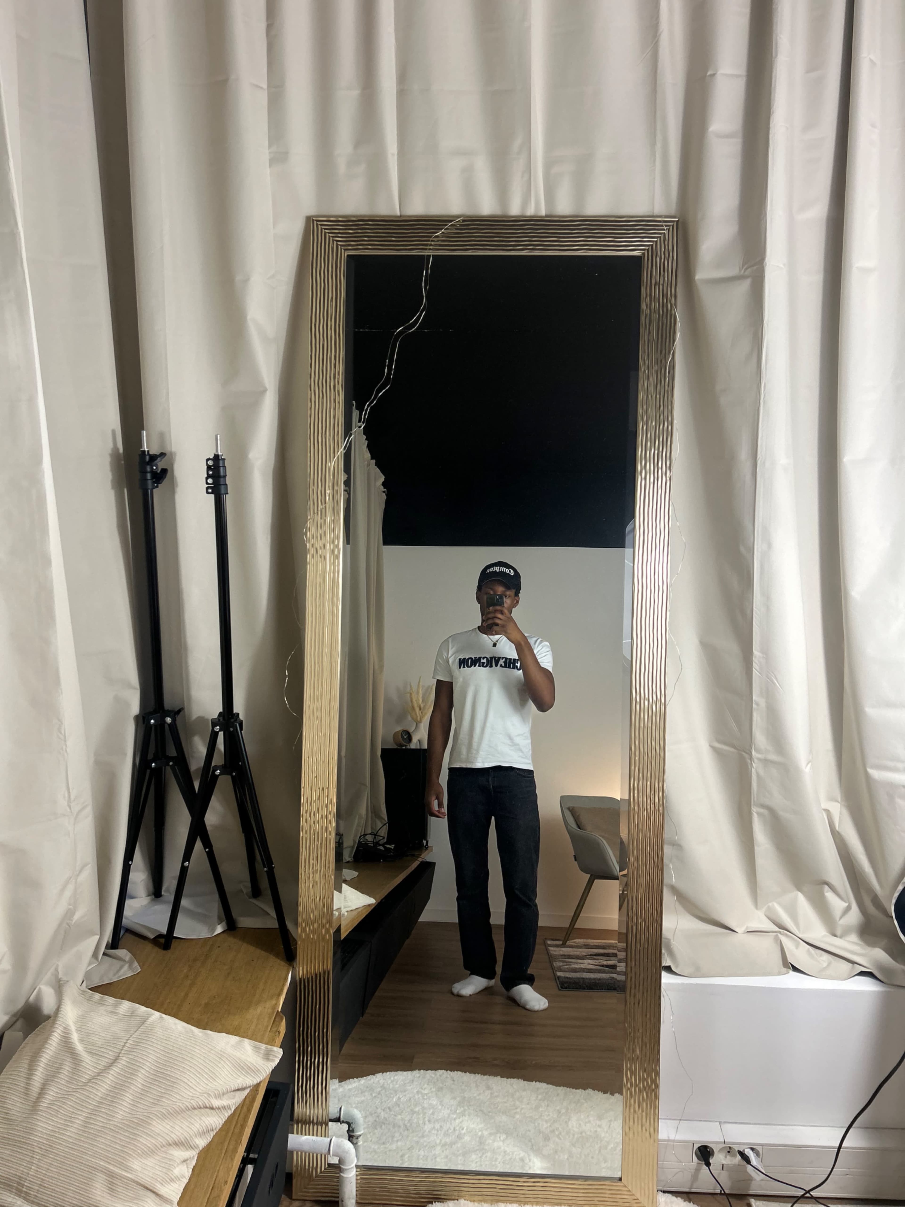 A person is standing in front of a tall mirror, wearing a white t-shirt and jeans, with a contemporary interior setting in the background.