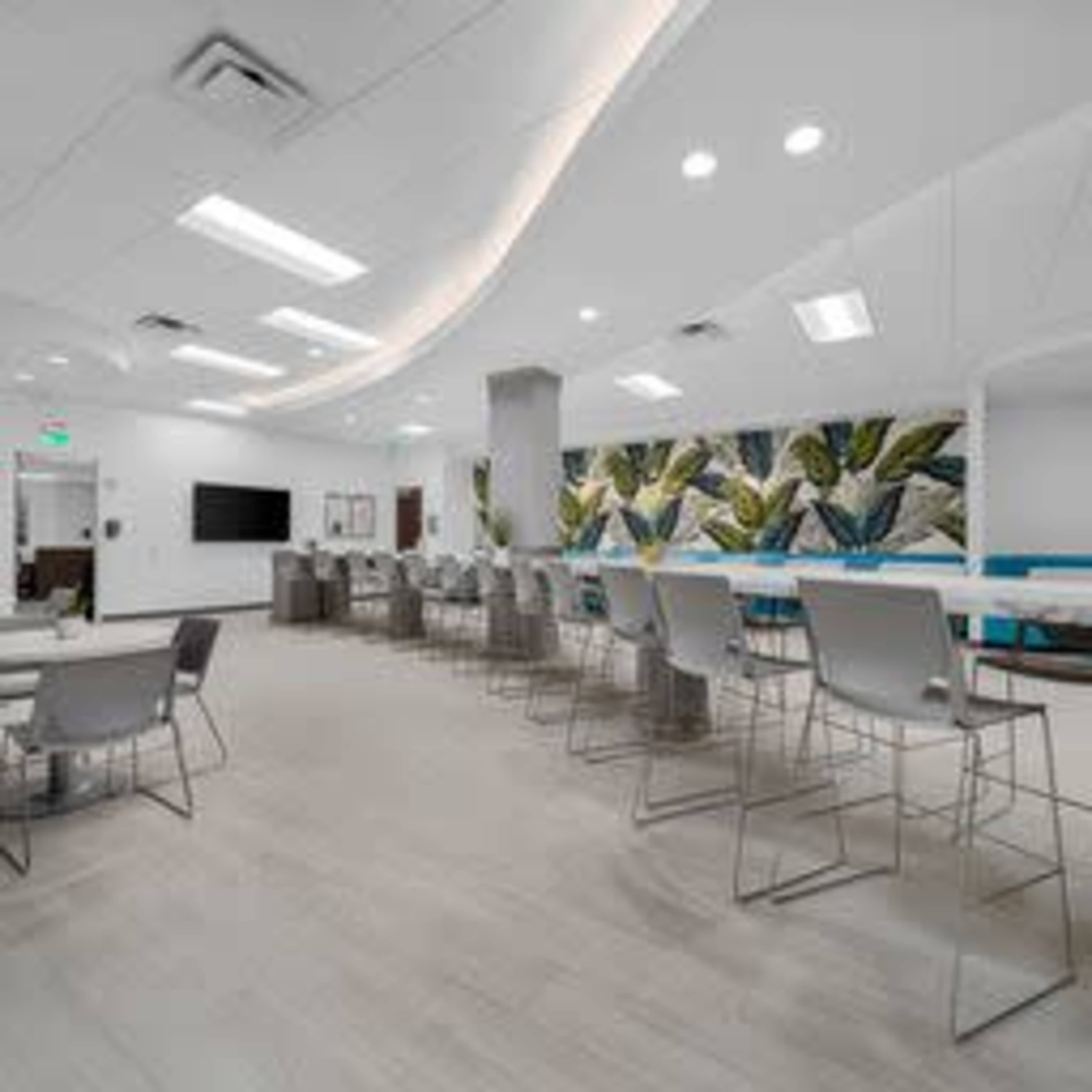 A modern, open office space with light-colored flooring, multiple seating areas, and a large mural of foliage on one wall.