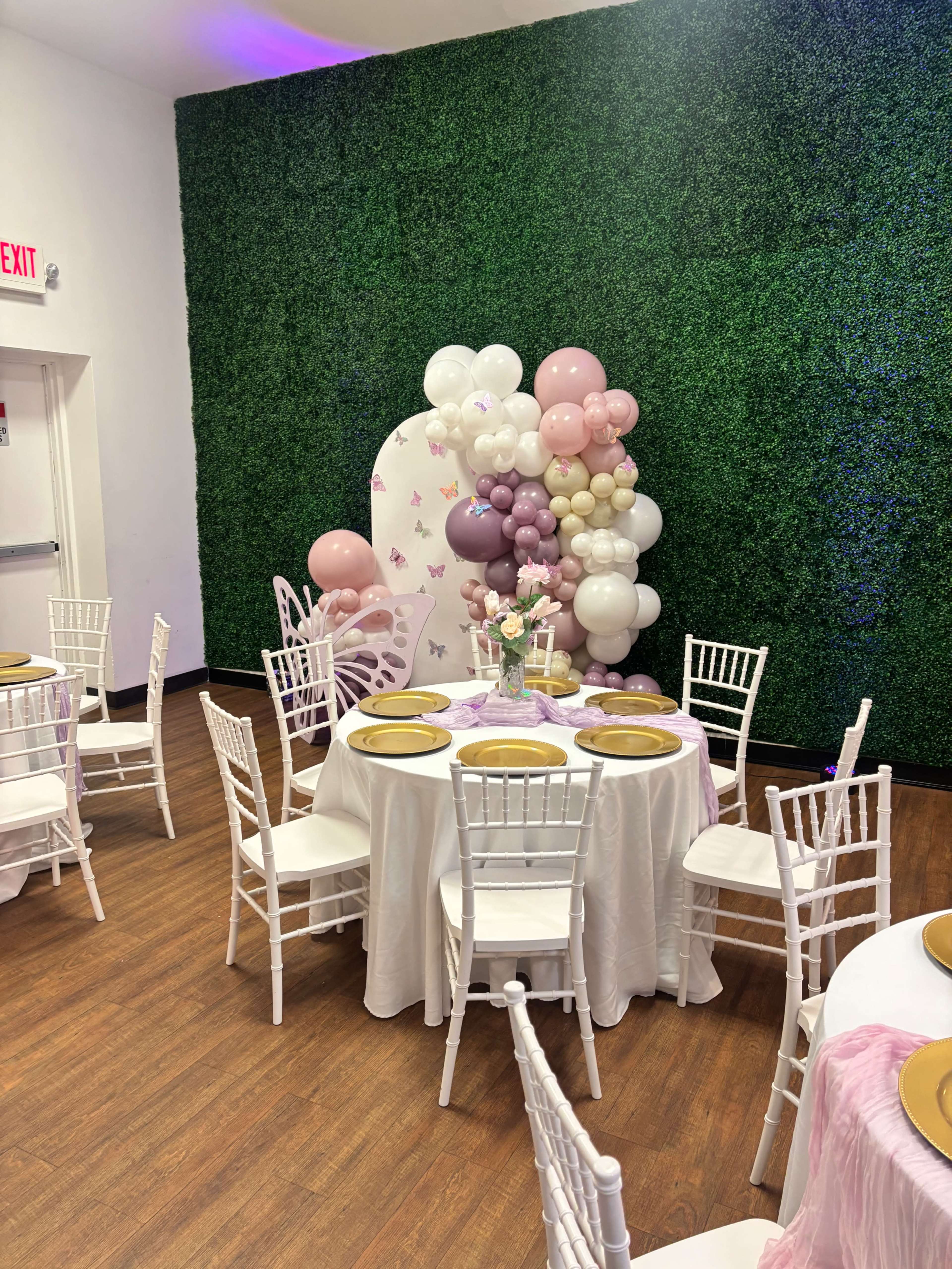The image shows a decorated event space with white tables set up, adorned with gold plates, and a vibrant balloon arrangement in shades of pink and purple against a green wall.