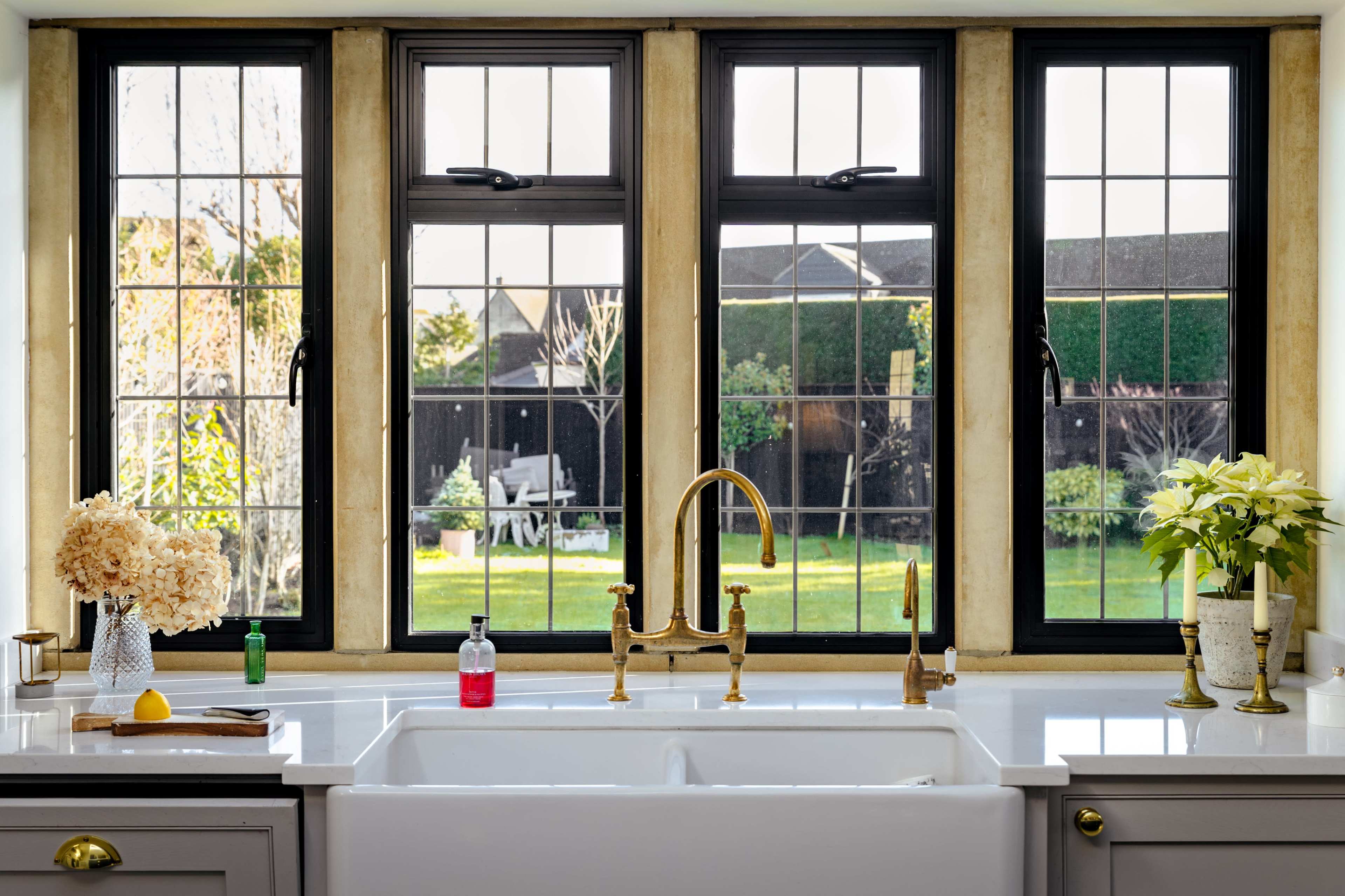 The image shows a modern kitchen sink with a large window above it, offering a view of a garden with greenery and outdoor furniture.