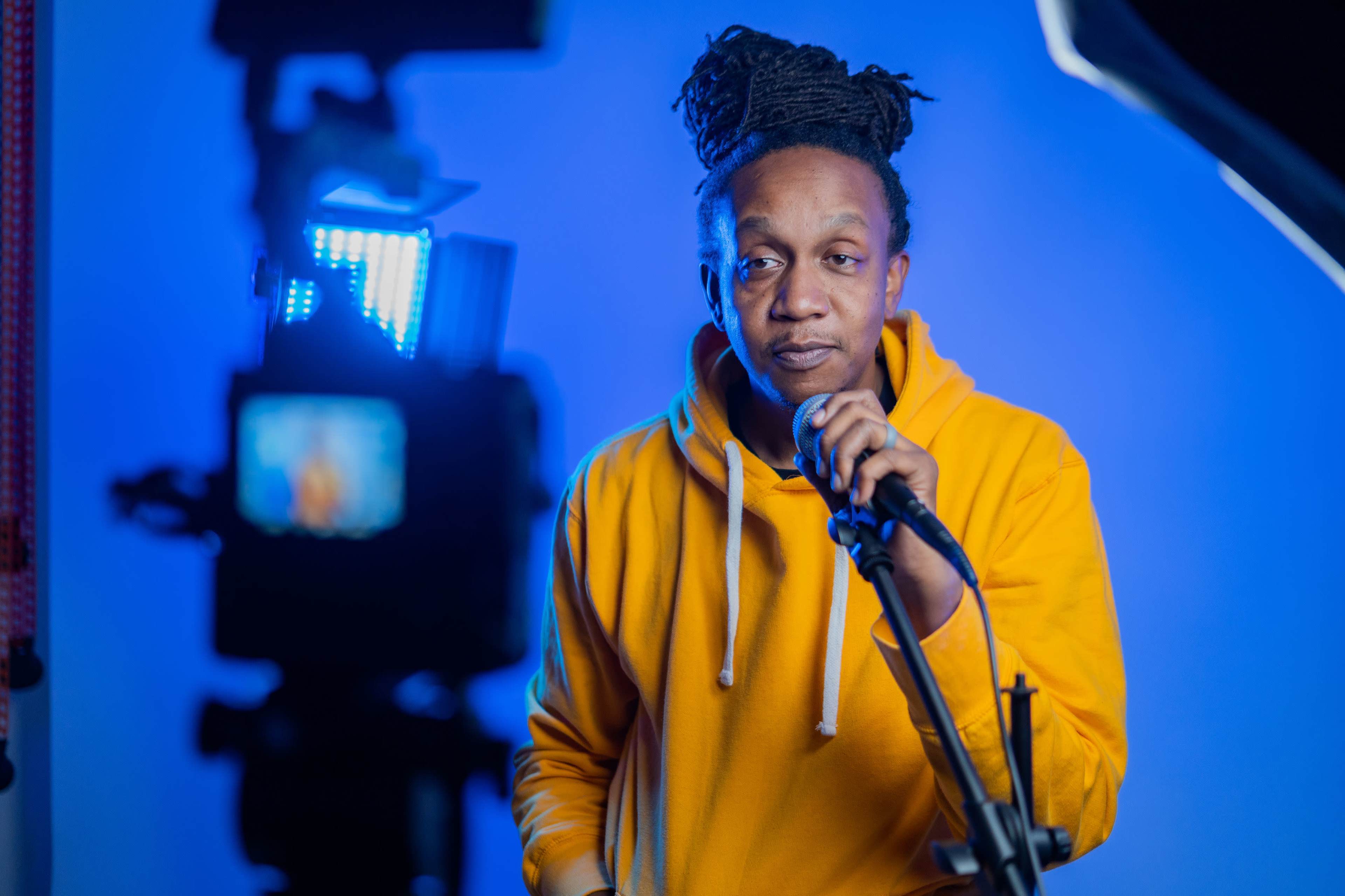 A person in a yellow hoodie stands in front of a microphone, speaking into it while being illuminated by a blue backdrop and studio lights.
