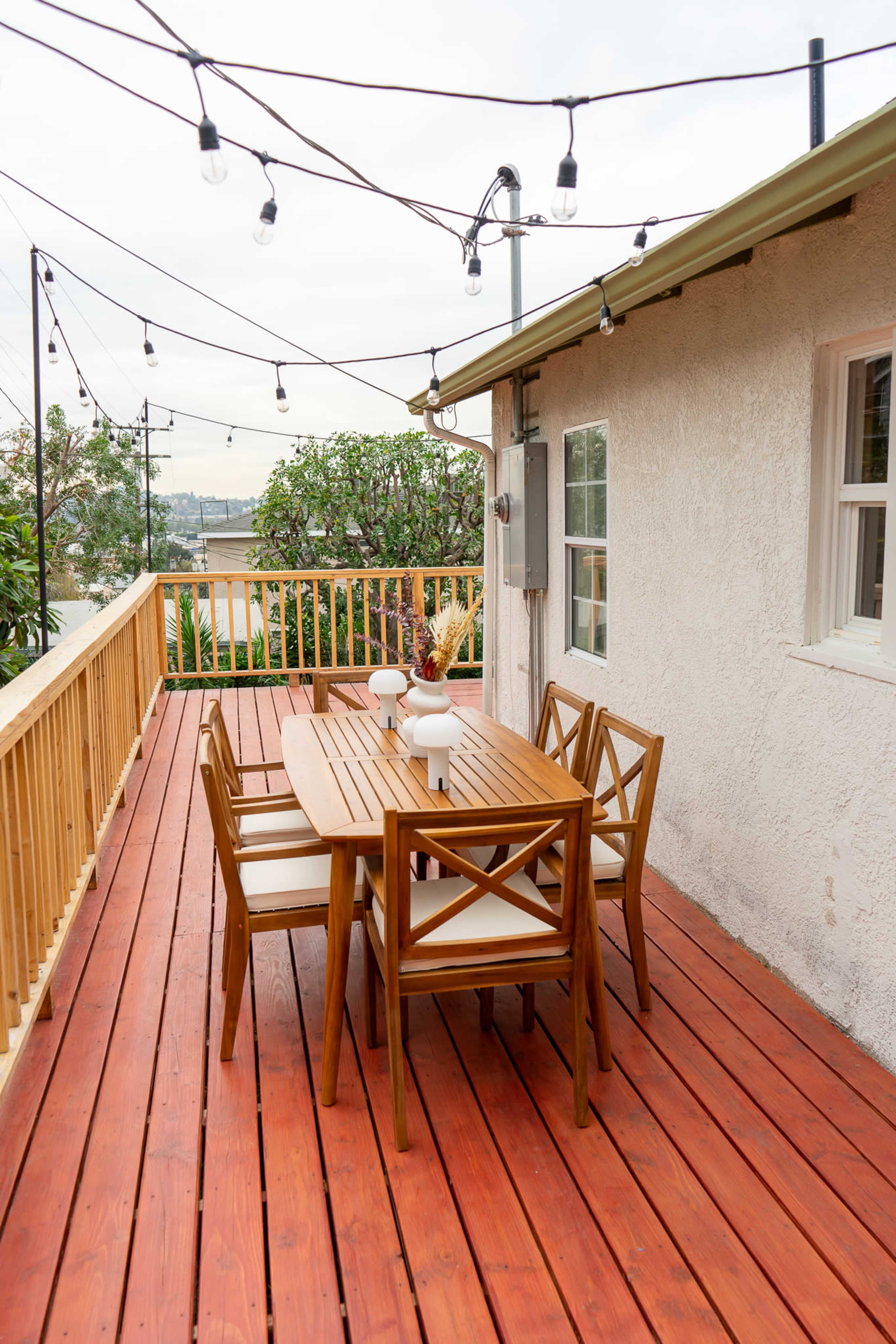 A wooden dining table with chairs is set up on a deck surrounded by string lights and a view of nearby trees.
