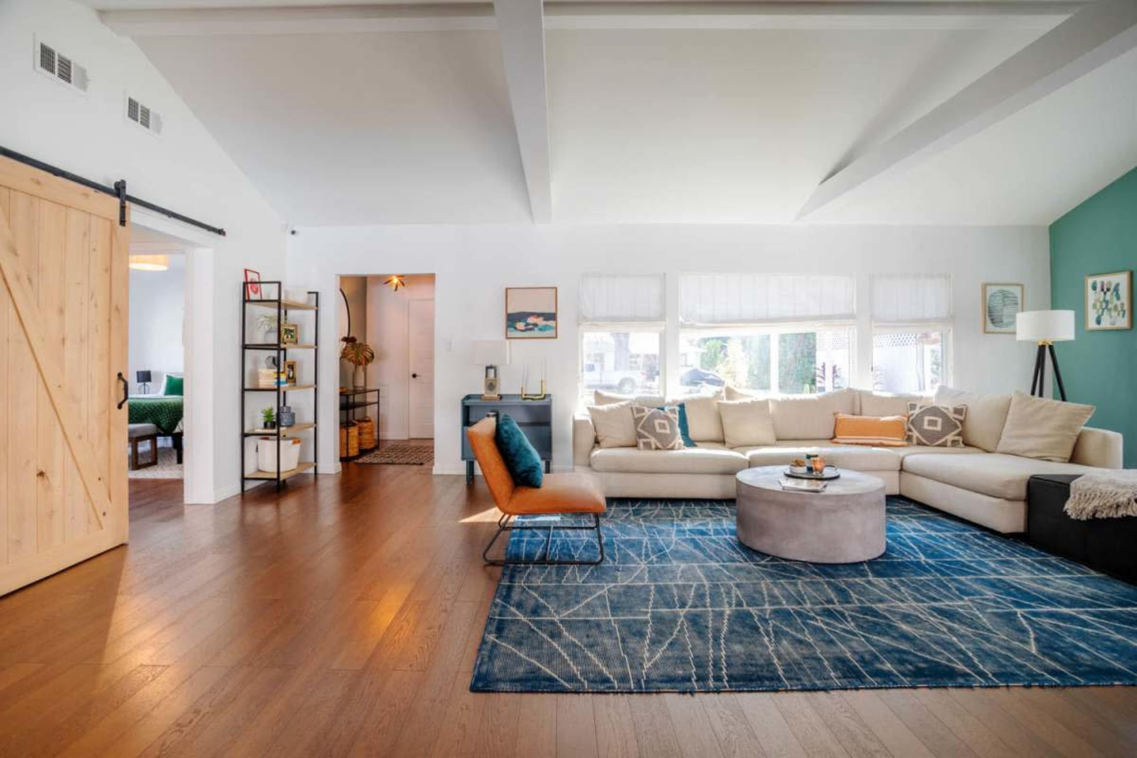 The image shows a modern living room with a beige sectional sofa, a round coffee table, and a blue area rug, featuring a wooden sliding door and a bookshelf in the background.