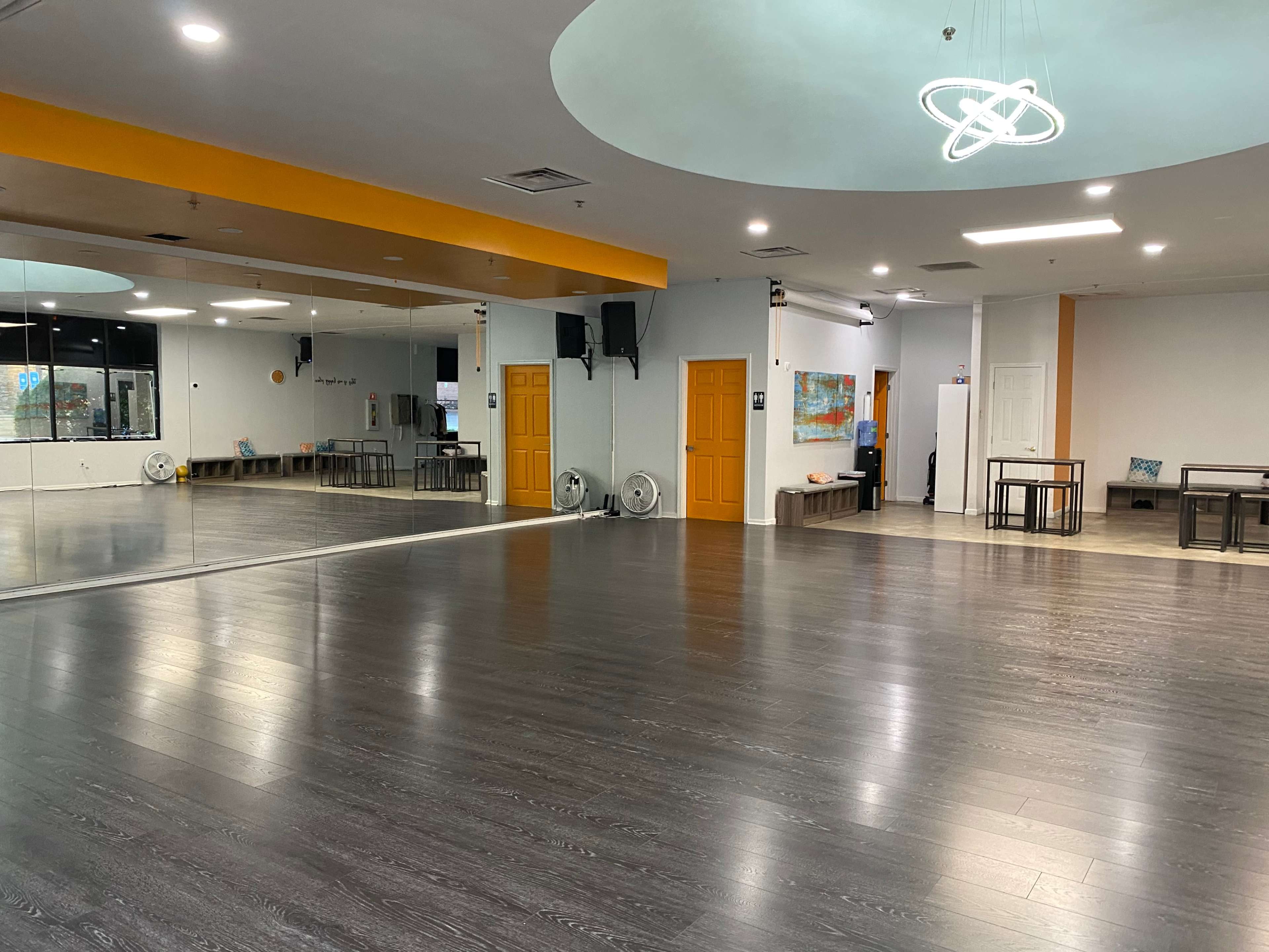 A spacious dance studio with wooden floors, large mirrors on one wall, and orange accents in the ceiling lighting.